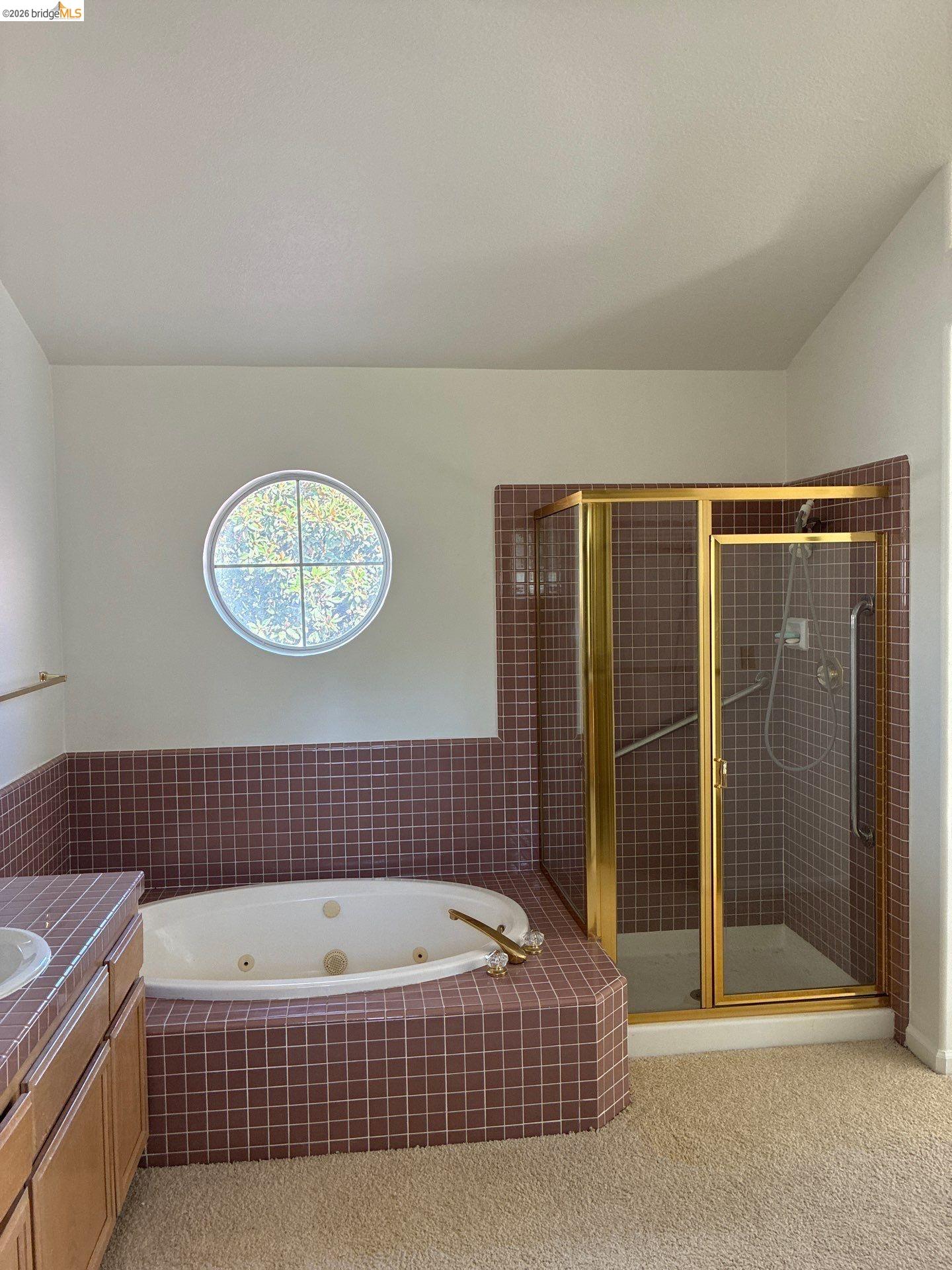 Jetted bathtub with tiled surround, separate shower with gold-tone frame, dual vanity with wood-finish cabinetry, and a circular window