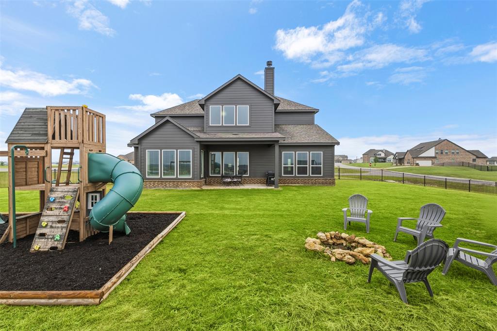 107 Yampa Court Rhome, TX 76078 - Photo 5 of 30 Expansive rear yard featuring a manicured lawn, established play structure with slide, and a stone-ringed fire pit area