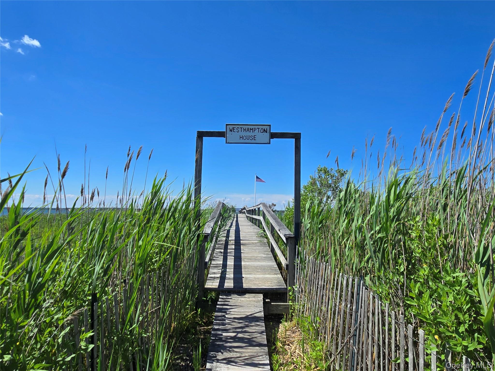 585 Dune Road, Unit L10 Westhampton, NY 11978 - Photo 23 of 25 Wooden boardwalk extending over water, flanked by tall green reeds and a rustic wooden fence