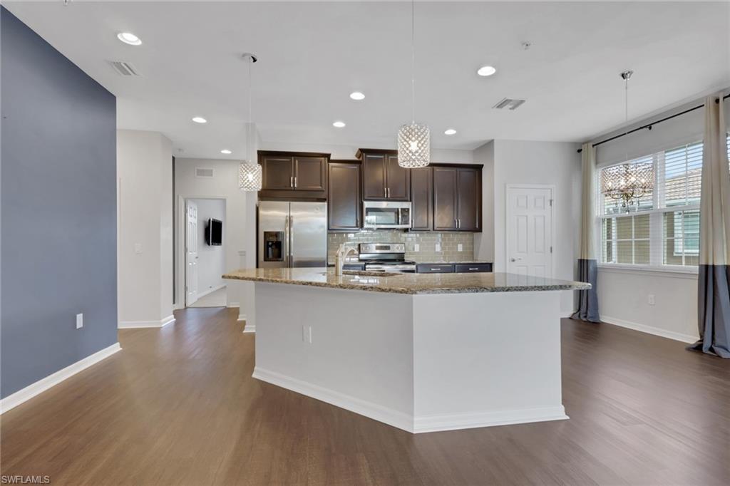 Kitchen featuring dark brown cabinetry, light stone counters, decorative light fixtures, appliances with stainless steel finishes, and a kitchen island with sink