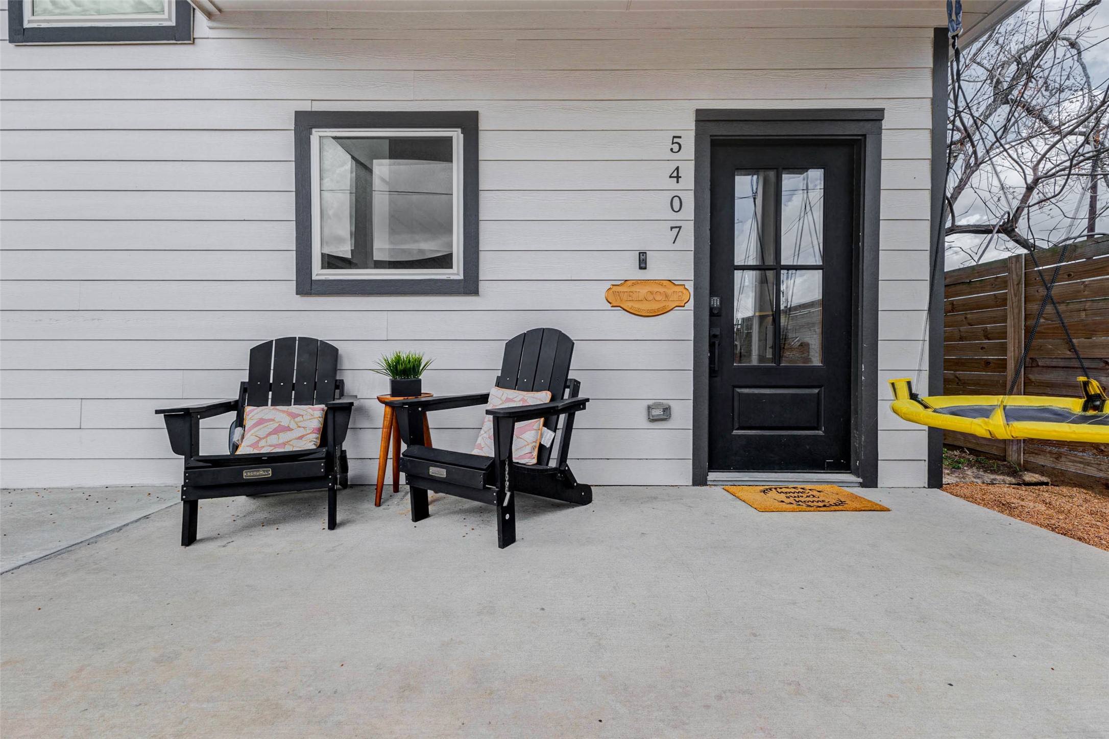 5407 Lee Street Houston, TX 77020 - Photo 16 of 17 Cozy front porch with two Adirondack chairs and cushions, perfect for relaxing. Features a small side table with a plant, a welcoming doormat, and a modern black door. The space is framed by horizontal siding and a wooden fence, providing a private, inviting entrance.