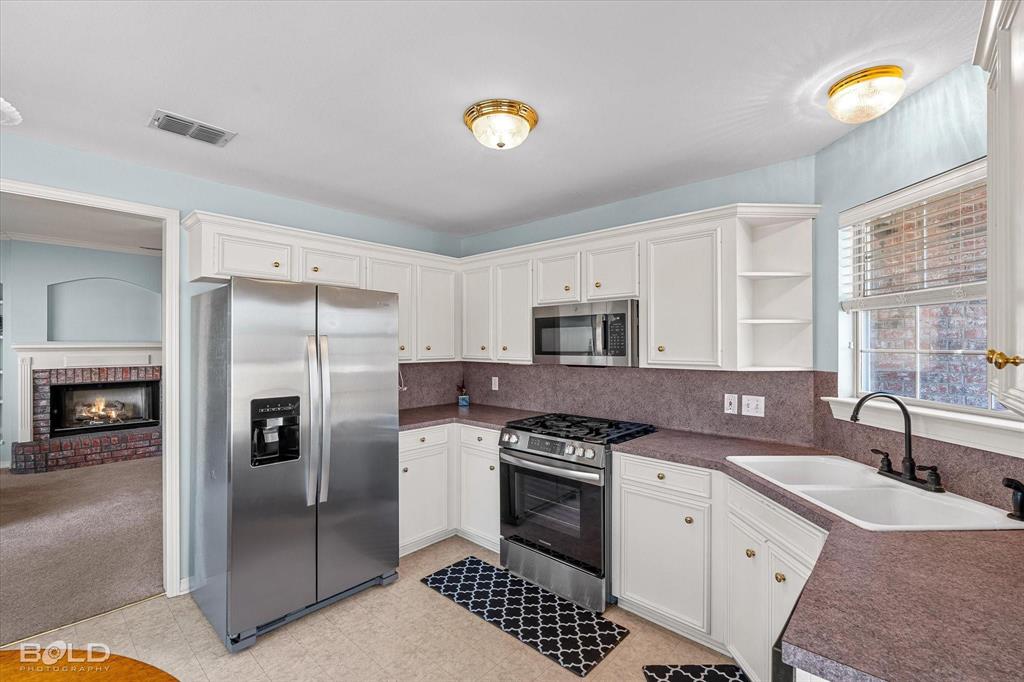 136 Scarborough Road Benton, LA 71006 - Photo 16 of 37 Kitchen featuring white cabinetry, stainless steel appliances, a corner sink with a black faucet, and a view into an adjacent room with a brick fireplace