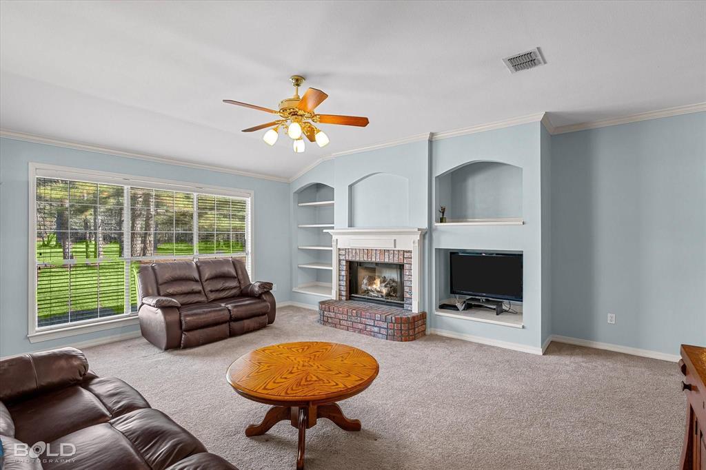 136 Scarborough Road Benton, LA 71006 - Photo 10 of 37 Living room featuring a brick fireplace with a white mantel, built-in shelving, a large window overlooking a treed landscape, a ceiling fan with integrated lighting, and neutral carpeting