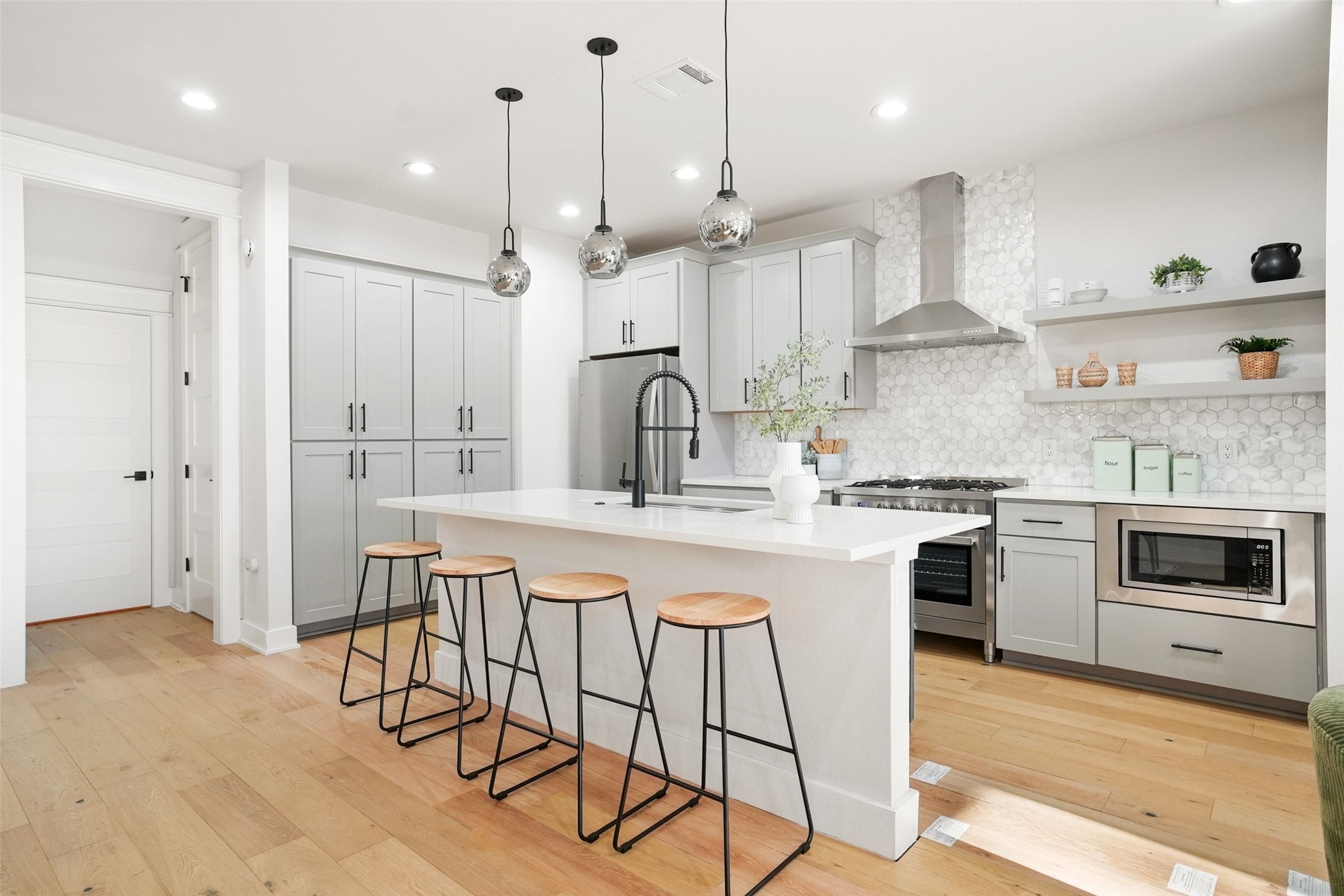 Modern kitchen featuring light wood-finish flooring, light grey cabinetry, a central island with white countertop, stainless steel appliances, and a hexagon tile backsplash