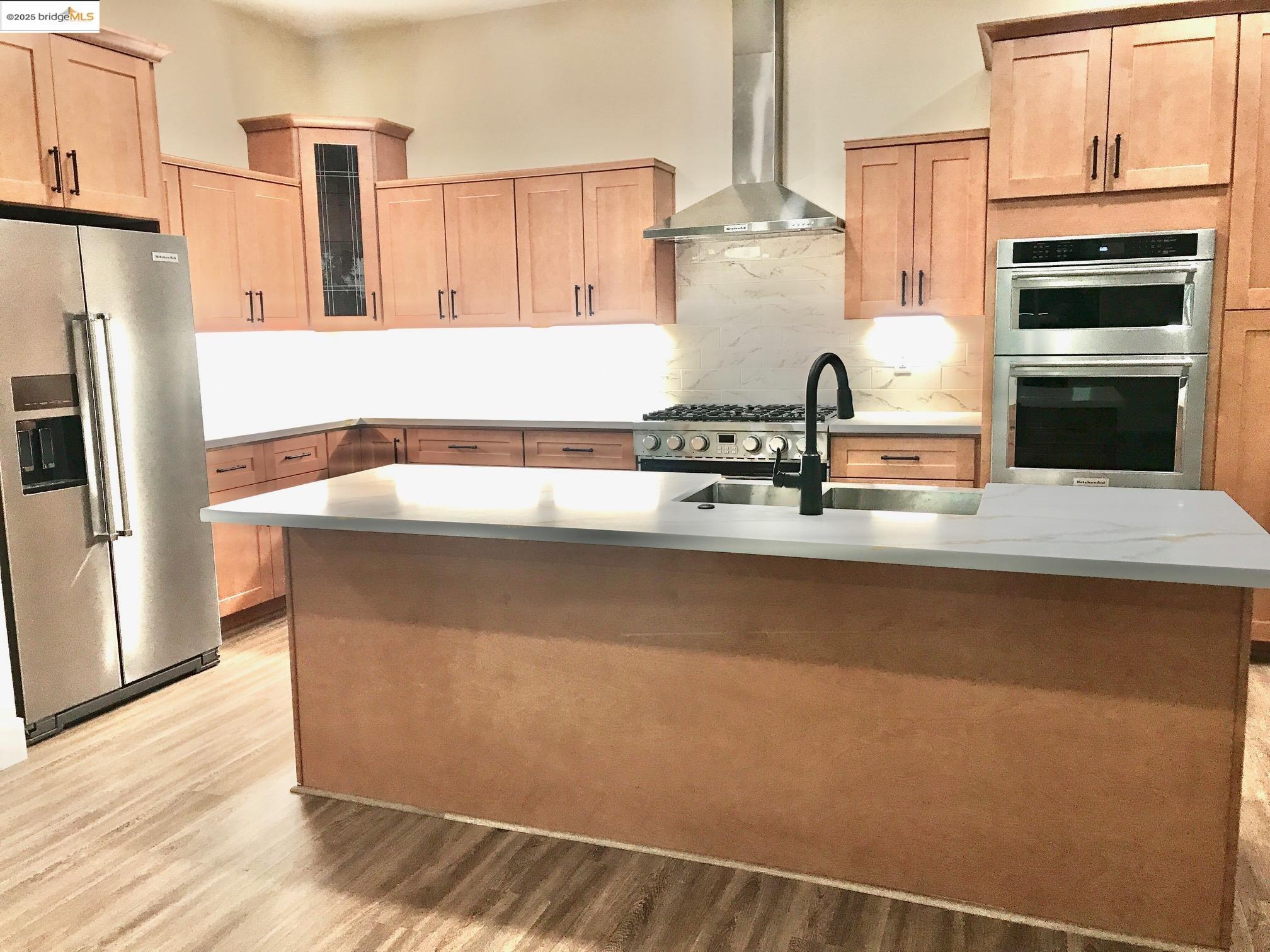 Kitchen with wall chimney range hood, appliances with stainless steel finishes, light brown cabinets, a kitchen island with sink, and light wood-style floors