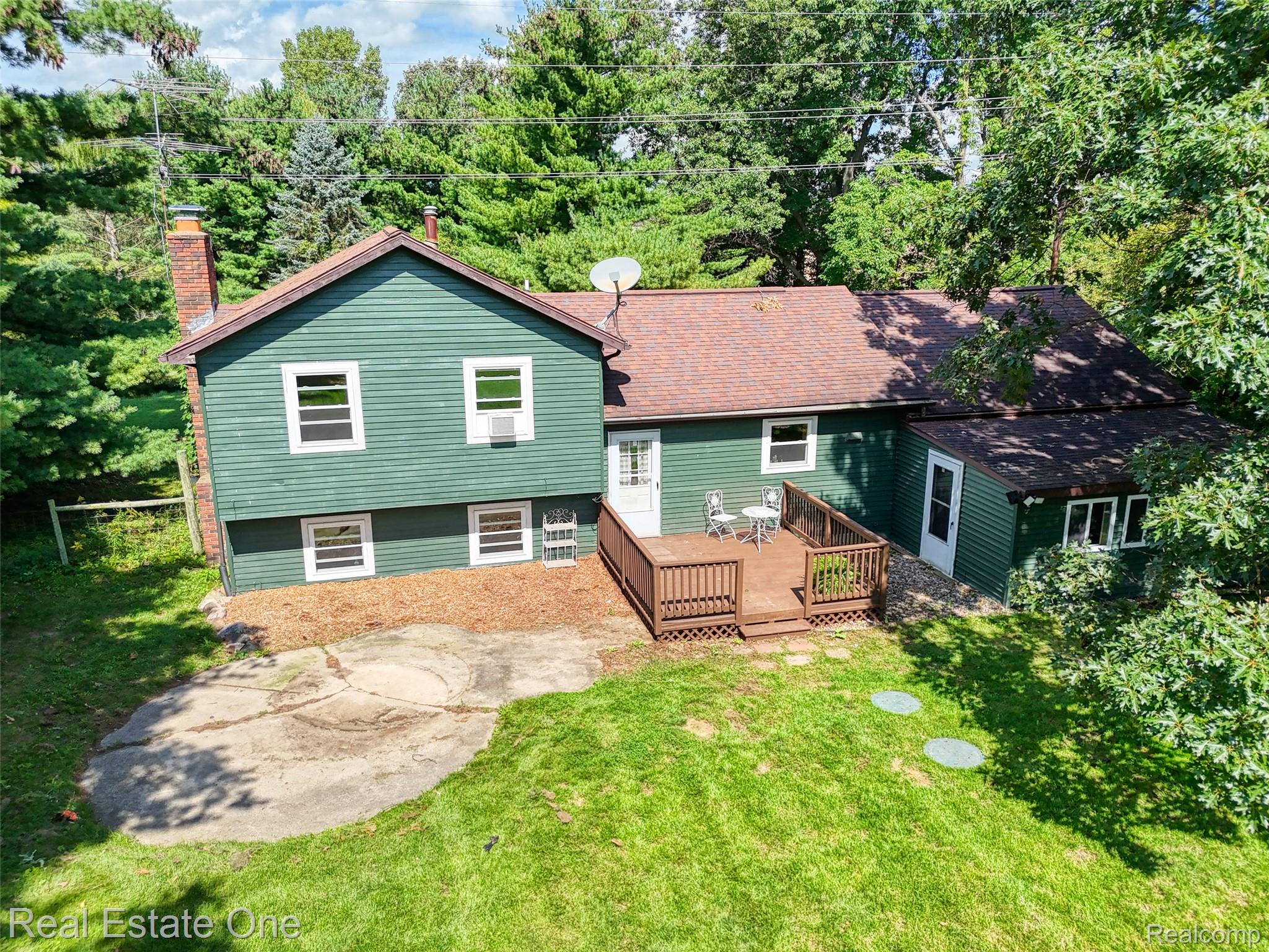 5130 Iosco Road Webberville, MI 48892 - Photo 37 of 39 Green exterior siding, wood deck, brick chimney, gabled roof, and mature perimeter trees