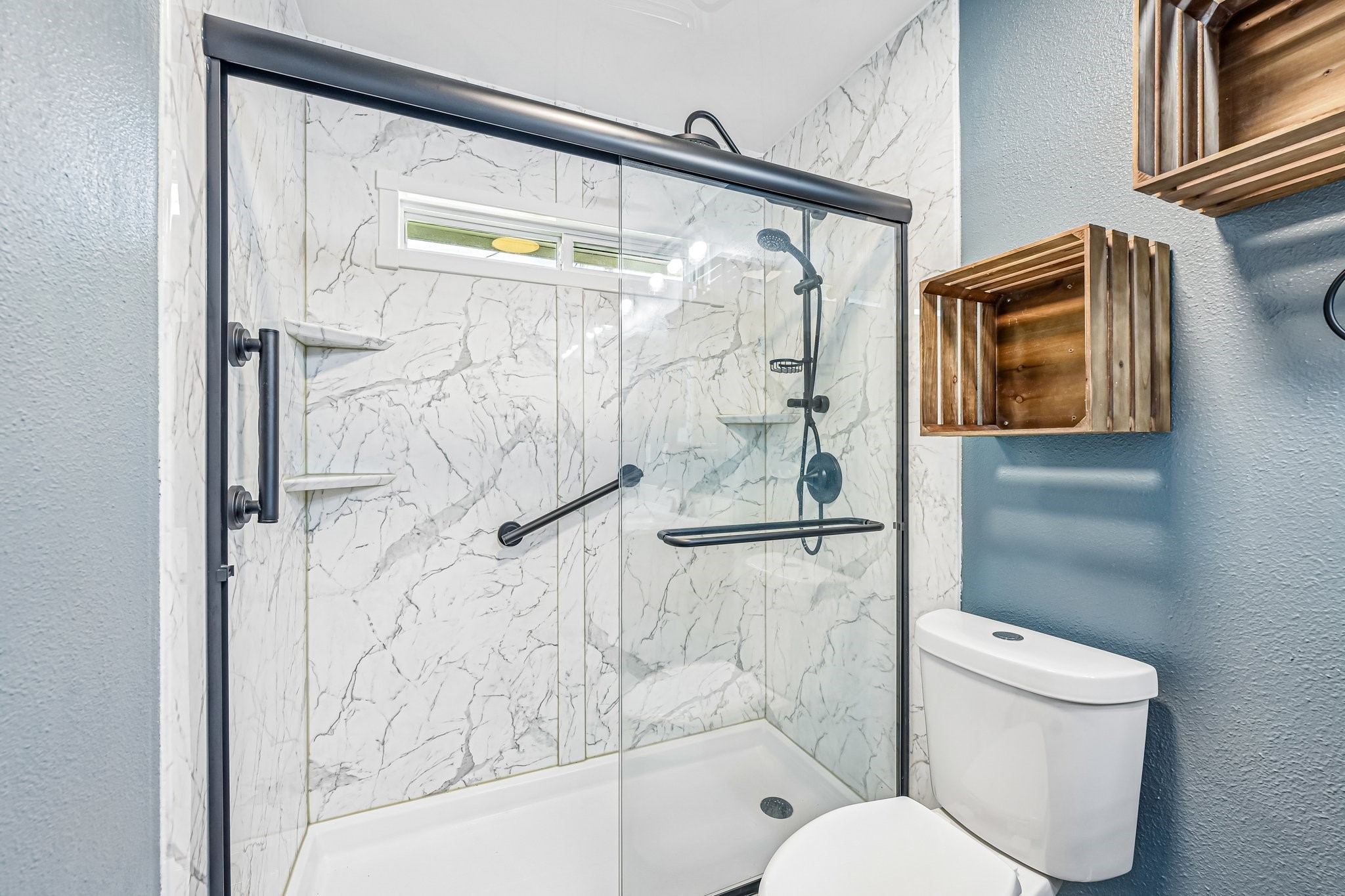 8 Arlington Street Conroe, TX 77301 - Photo 22 of 37 Modern bathroom featuring a sleek glass shower with elegant marble-patterned tiles, a stylish black showerhead, and convenient built-in shelving. The space is complemented by a white toilet and rustic wooden wall shelves, creating a contemporary yet cozy feel.