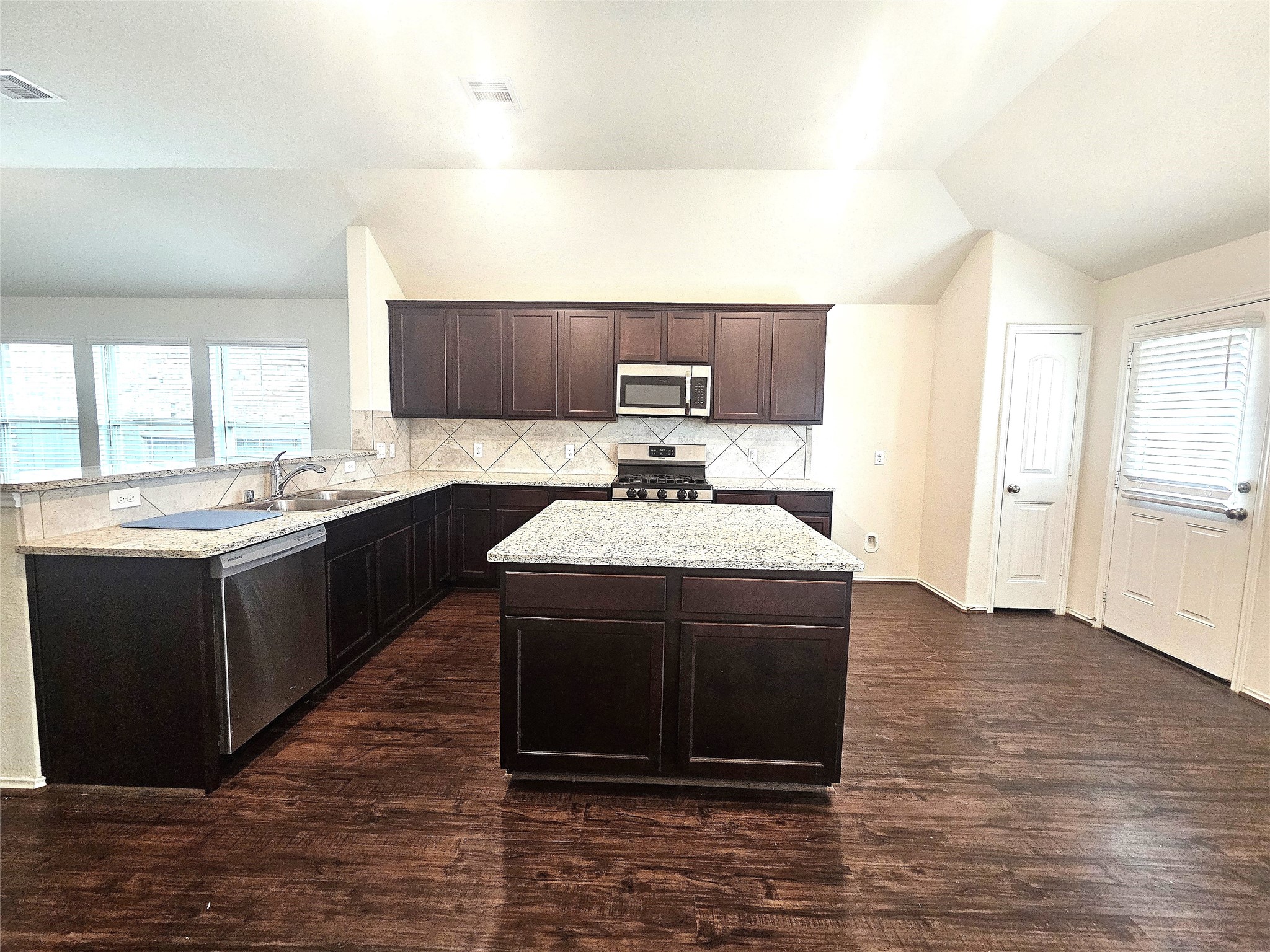17439 Chester Valley Trail Hockley, TX 77447 - Photo 8 of 22 This kitchen features dark wood cabinets, a central island with granite countertops, stainless steel appliances, and tile backsplash. It has wood flooring, ample natural light from windows, and a door leading outside.