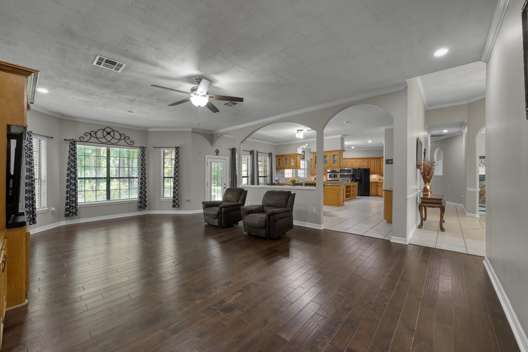 910 Commons Way Court Huffman, TX 77336 - Photo 8 of 49 This spacious living area features hardwood floors, large windows with decorative curtains, and a ceiling fan. It opens to a tiled kitchen with wooden cabinetry, creating a welcoming and functional space.