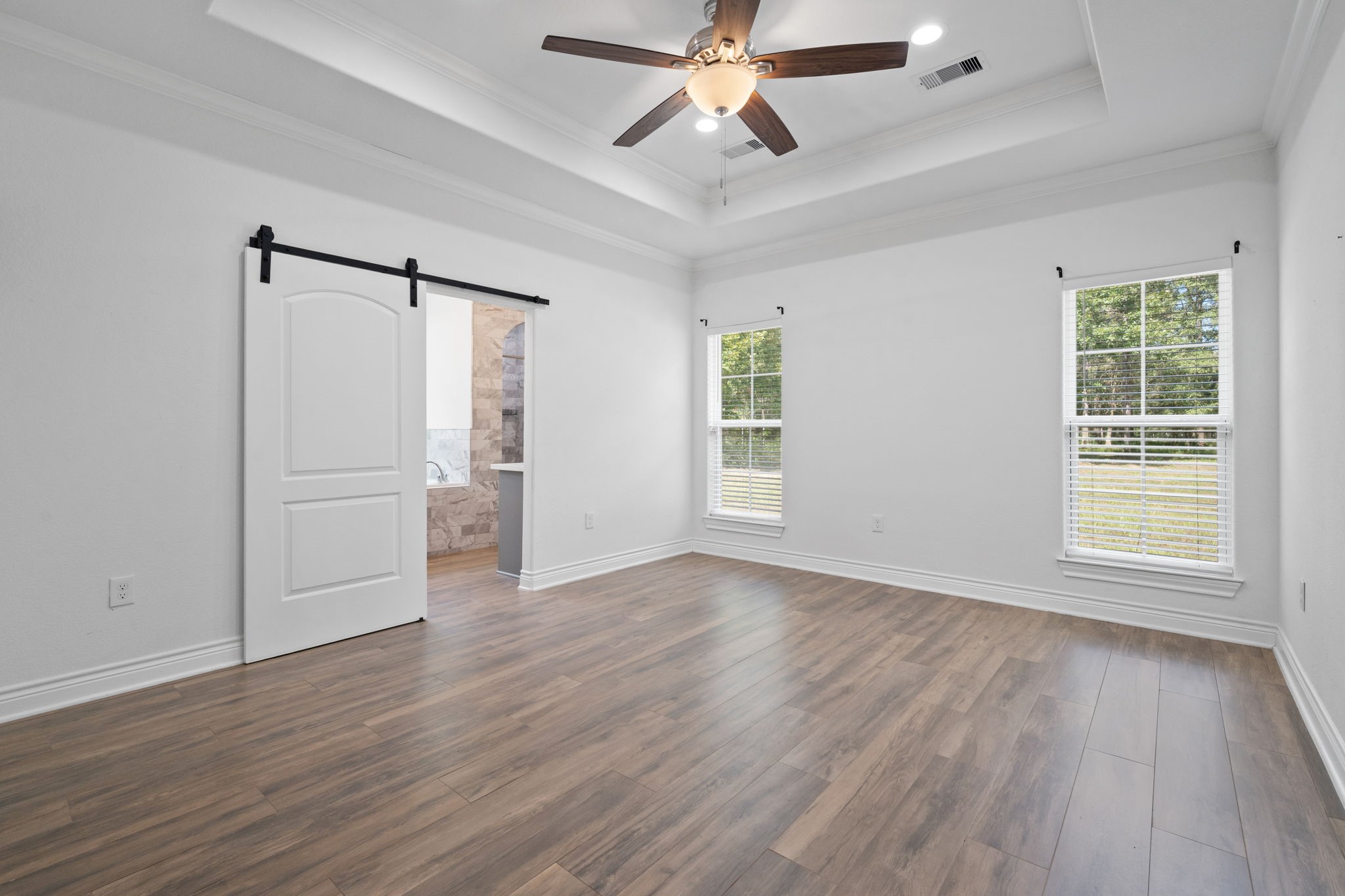 7815 Timber Switch Road Cleveland, TX 77328 - Photo 26 of 39 Spacious bedroom featuring elegant wood flooring, a charming tray ceiling with a ceiling fan, and abundant natural light from dual windows. A stylish sliding barn door leads to the en-suite bathroom.