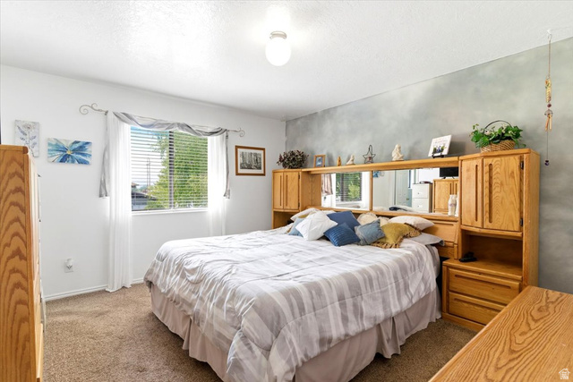 5321 South 3100th Street West Roy, UT 84067 - Photo 40 of 64 Carpeted bedroom featuring a large window with blinds, a prominent accent wall, and a built-in wood-finish headboard unit with integrated storage and mirrored panels