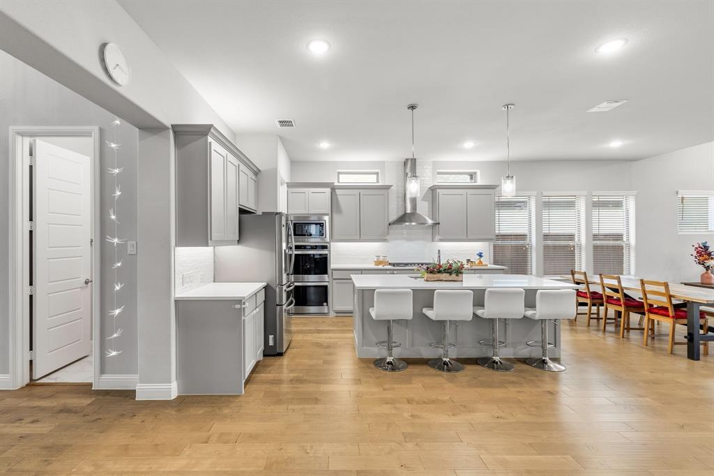 7852 Buffaloberry Road Frisco, TX 75035 - Photo 5 of 36 Open-concept kitchen featuring an oversized island with pendant lighting, light wood-finish flooring, stainless steel appliances, grey shaker cabinetry, and a white subway tile backsplash