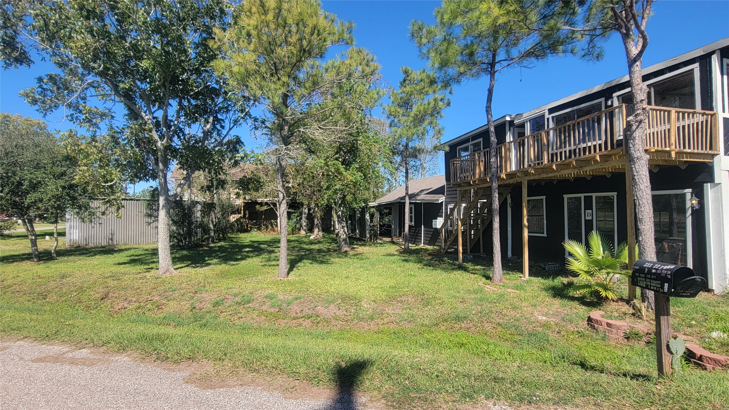 12018 Sandpiper Road Angleton, TX 77515 - Photo 44 of 48 This photo shows a two-story home with a rustic wooden exterior and a balcony. The property is surrounded by trees, providing a natural and serene setting. A well-maintained grassy yard leads to the front entrance, and a mailbox is visible near the road.