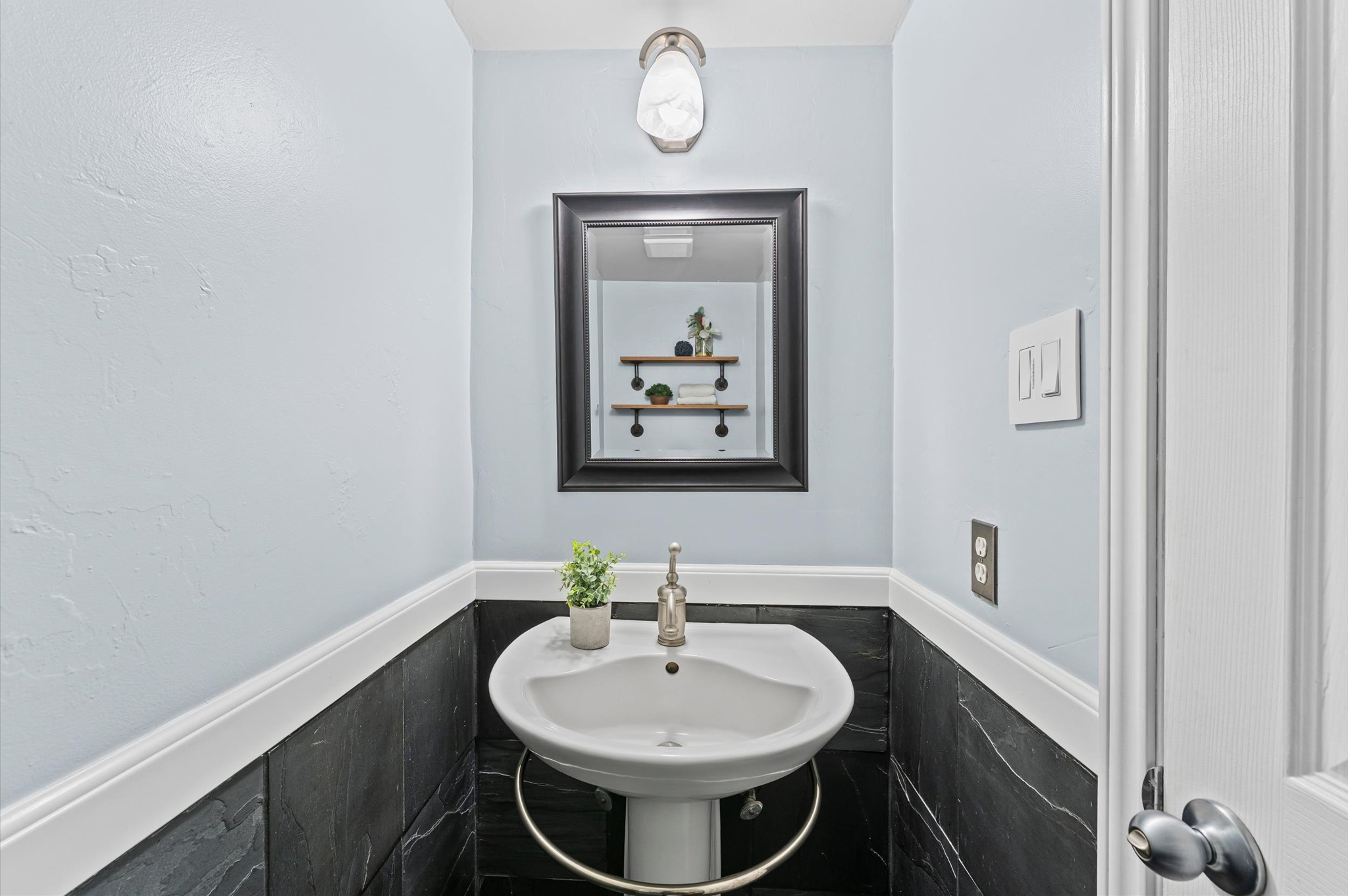 9810 Charter Ridge Drive Houston, TX 77070 - Photo 8 of 28 This is a compact, modern powder room featuring light blue walls, a pedestal sink, and a framed mirror. It has a stylish light fixture and a decorative plant for a touch of elegance.