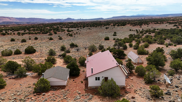 358 South Sky Drive Torrey, UT 84775 - Photo 5 of 82 White exterior residence with a red metal roof, accompanied by two smaller outbuildings featuring gray roofs, all set within a vast desert landscape with distant mountains