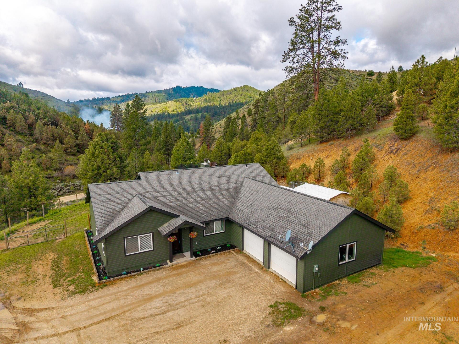4 Round Up Circle Boise, ID 83716 - Photo 2 of 47 Expansive property featuring a green-sided residence with a dark gray shingle roof, integrated two-car garage with white doors, surrounding hillsides with mature trees, and a small outbuilding