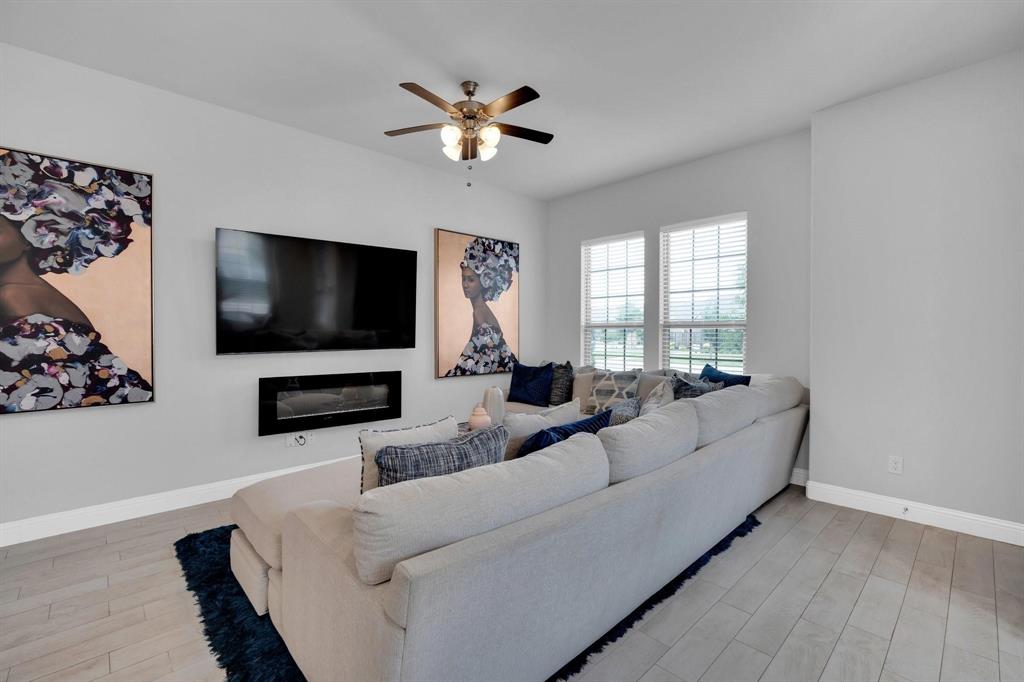 1498 Weston Road Dallas, TX 75234 - Photo 5 of 24 Bright living space featuring light gray walls, wood-finish flooring, two large windows with blinds, a wall-mounted fireplace, and a ceiling fan with integrated lighting