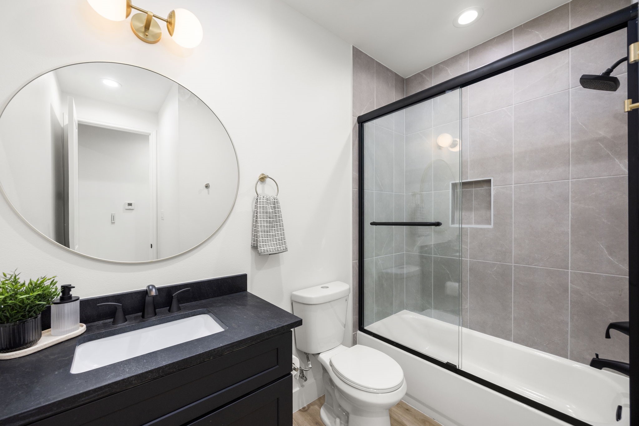 4611 Dewberry Street, Unit C Houston, TX 77021 - Photo 27 of 34 This modern bathroom features a sleek black vanity with a round mirror and double light fixture. It includes a glass-enclosed tub with elegant tile, a toilet, and light wood flooring, creating a clean and stylish space.