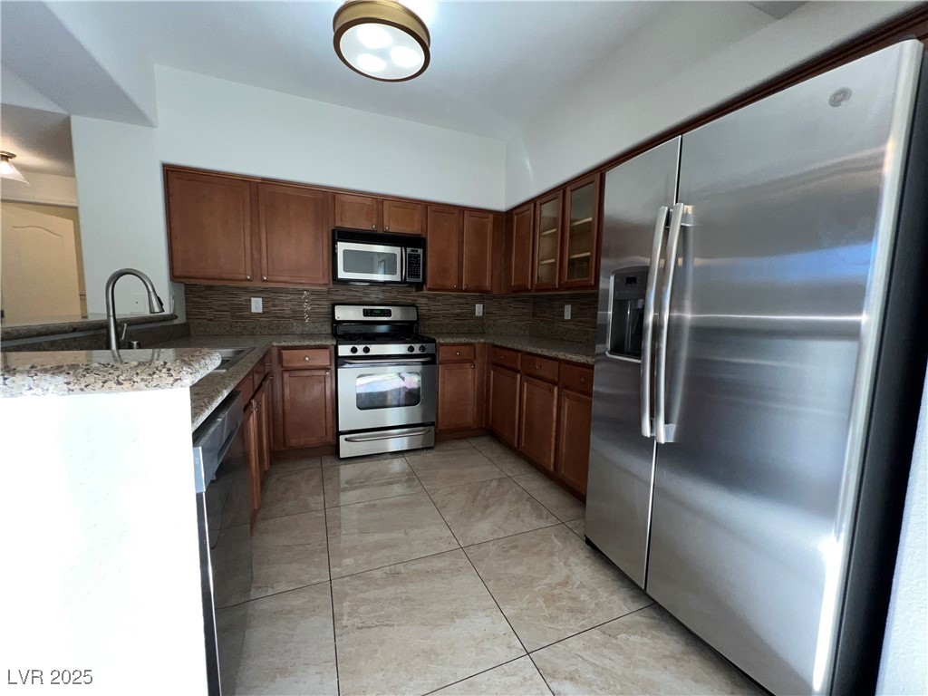 1705 King James Street, Unit 203 Las Vegas, NV 89144 - Photo 7 of 21 Kitchen featuring appliances with stainless steel finishes, glass insert cabinets, decorative backsplash, dark stone counters, and brown cabinets