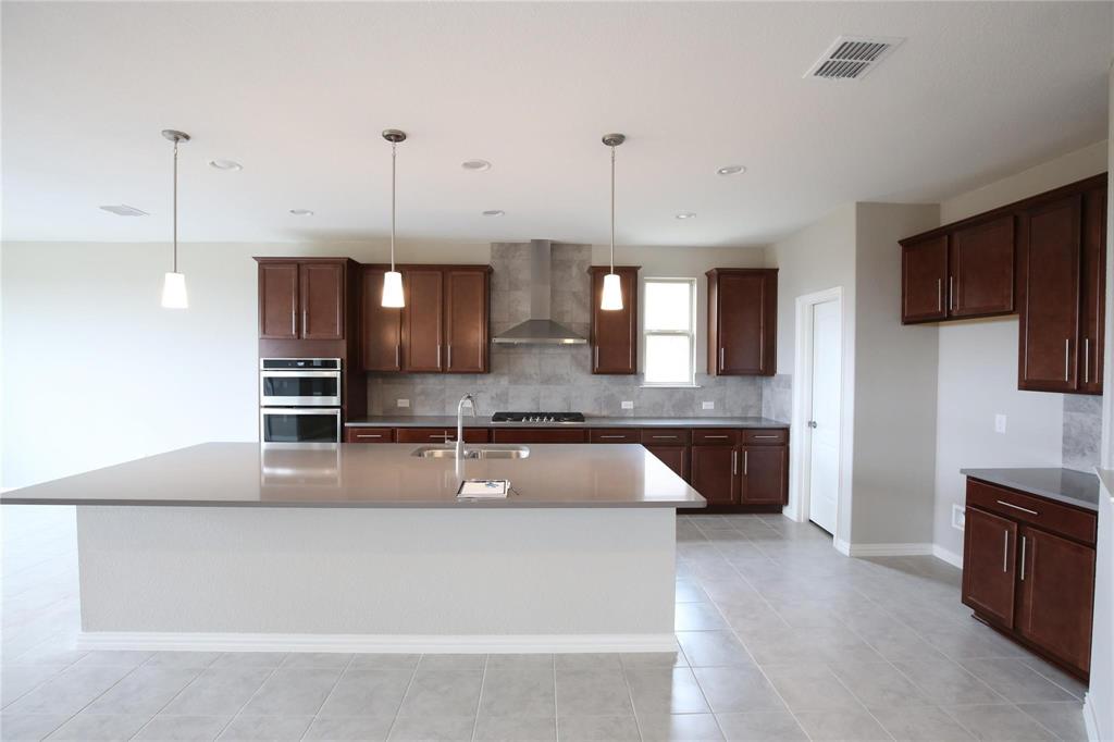 15142 Rosario Road Frisco, TX 75035 - Photo 4 of 21 Spacious kitchen featuring an expansive central island with an undermount sink, dark wood-finish cabinetry, built-in double ovens, and stainless steel range hood