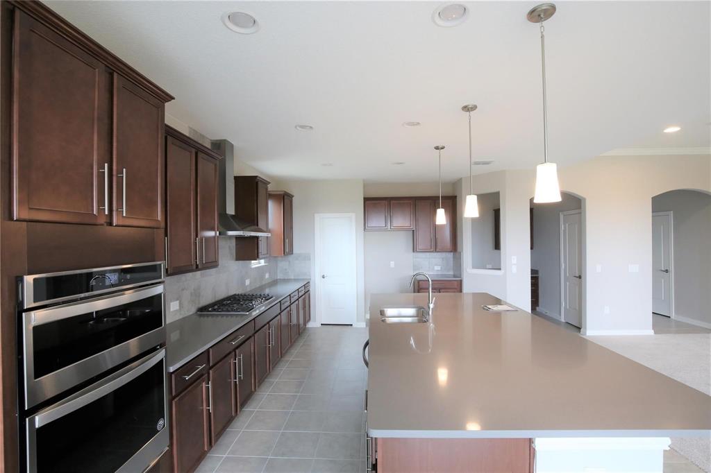 15142 Rosario Road Frisco, TX 75035 - Photo 6 of 21 Spacious kitchen featuring an expansive island with a built-in sink, pendant lighting, stainless steel double wall ovens, a gas cooktop with a range hood, and extensive dark wood cabinetry