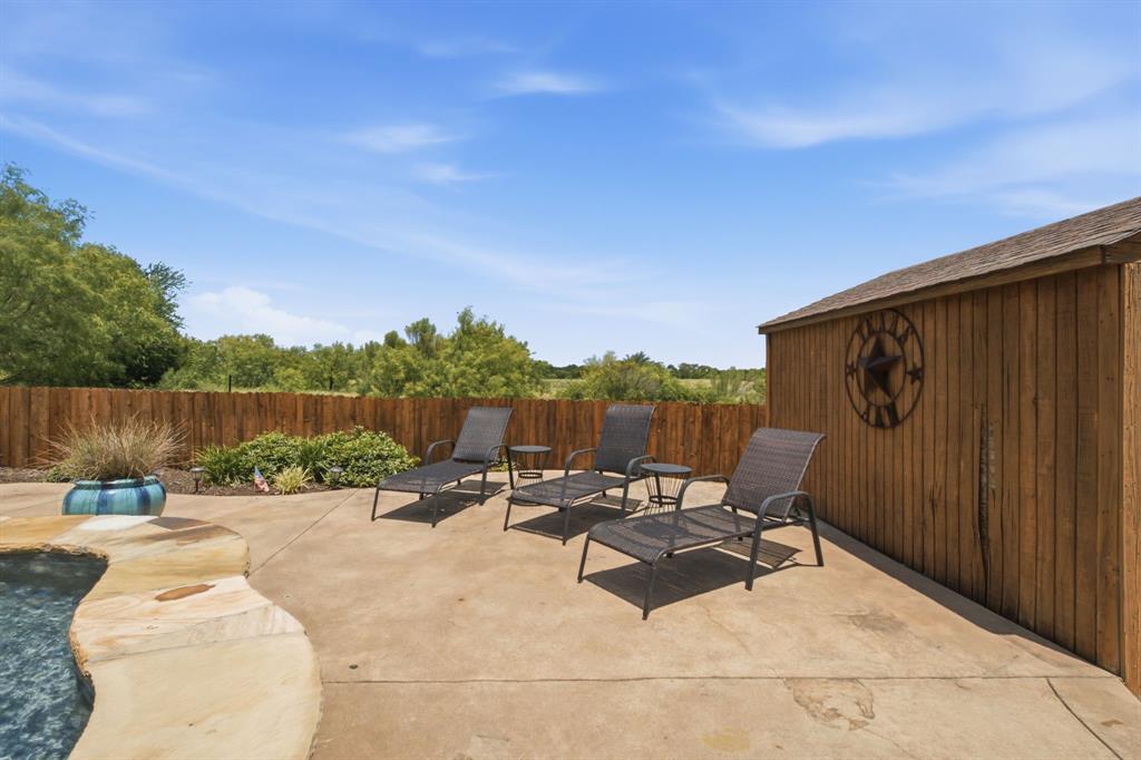 600 Aspen Street Pilot Point, TX 76258 - Photo 34 of 39 Expansive concrete patio with a natural stone-edged pool, wood privacy fence, and a matching wood-sided shed