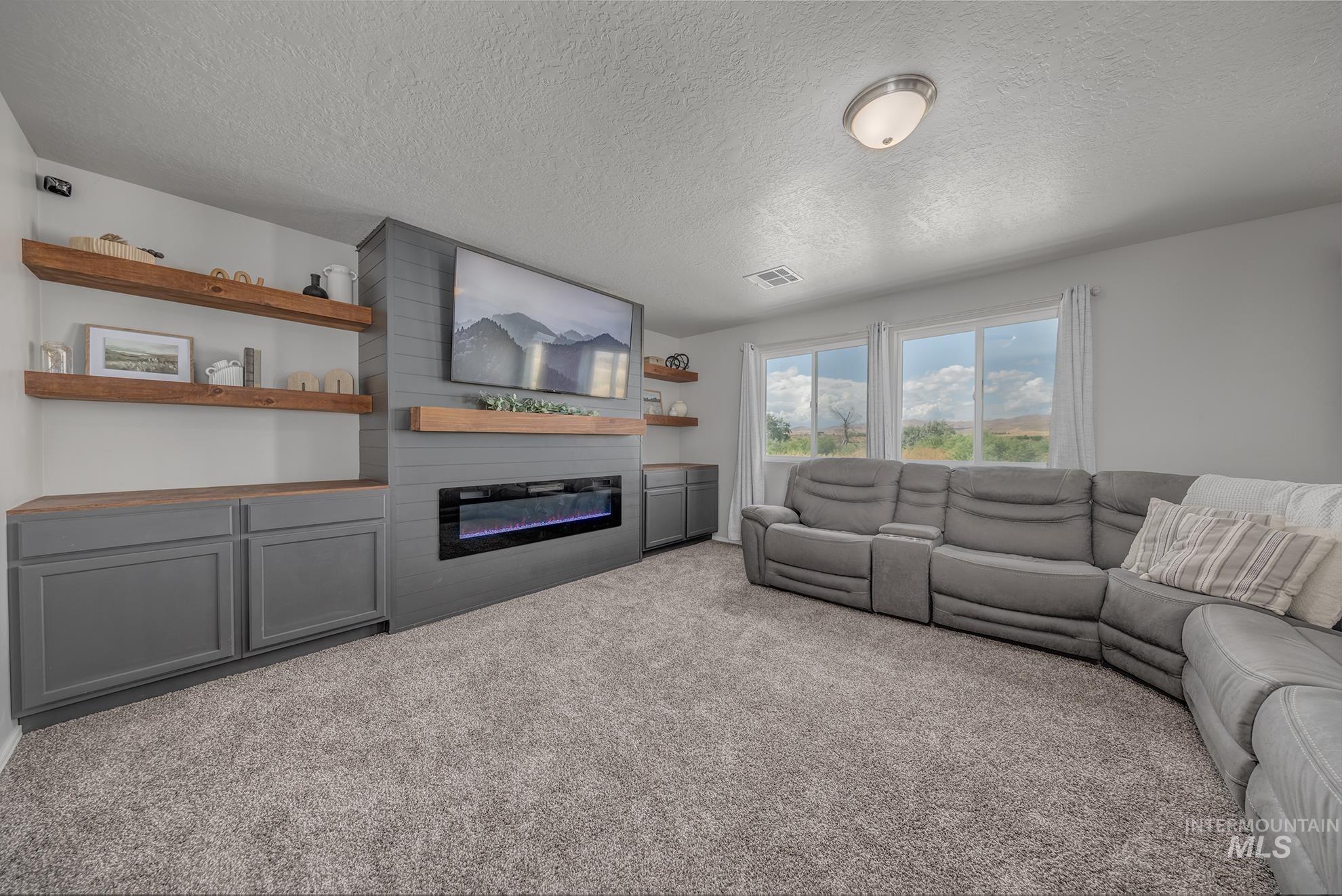 1049 Price Road Weiser, ID 83672 - Photo 21 of 47 Living area featuring a textured carpet floor, a built-in media center with grey cabinetry and wood-finish countertops, floating wood shelves, and a linear electric fireplace