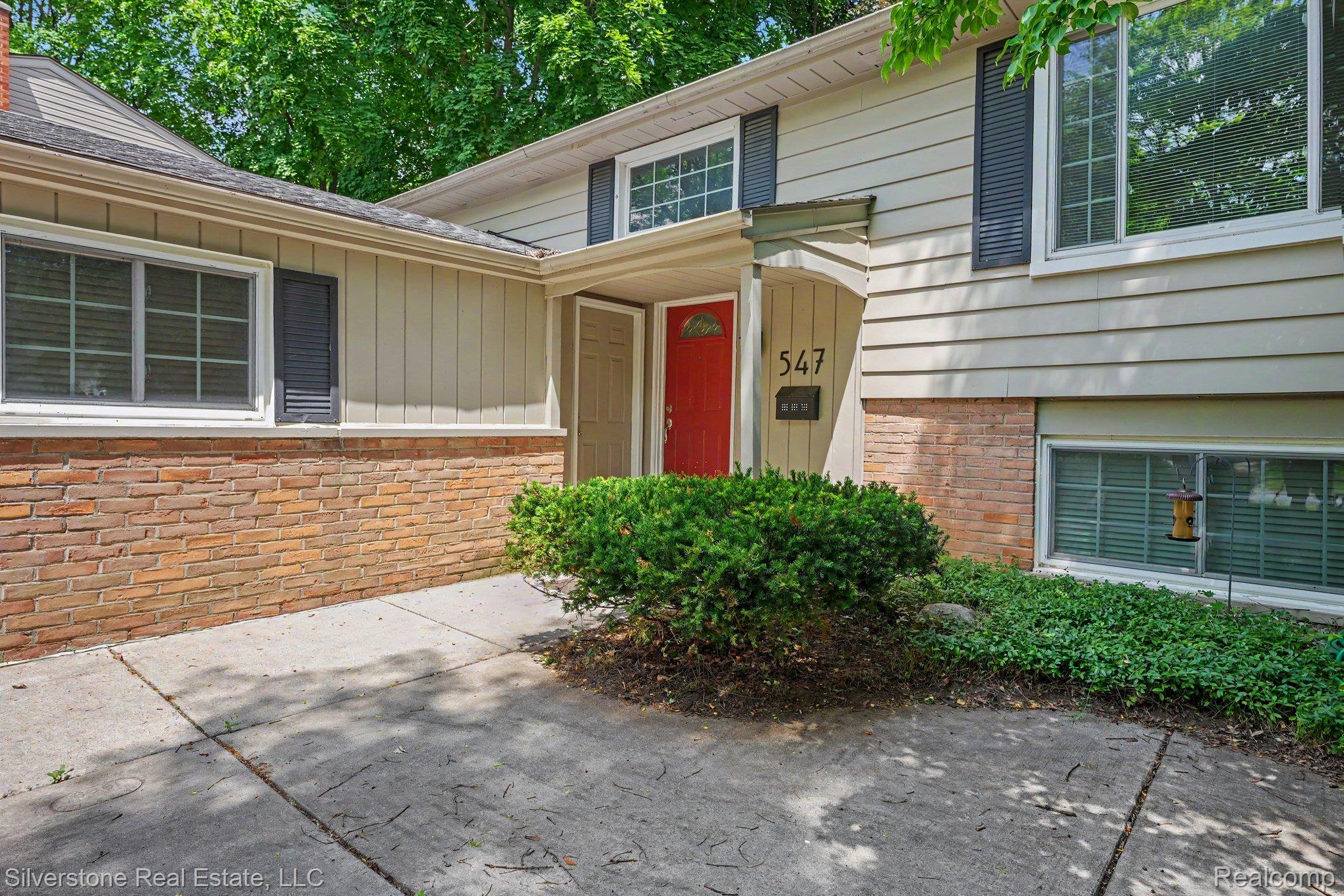 547 Reed Street Northville, MI 48167 - Photo 4 of 38 Mid-century exterior featuring light-colored siding, brick accent detailing, a vibrant red entry door, and dark louvered shutters