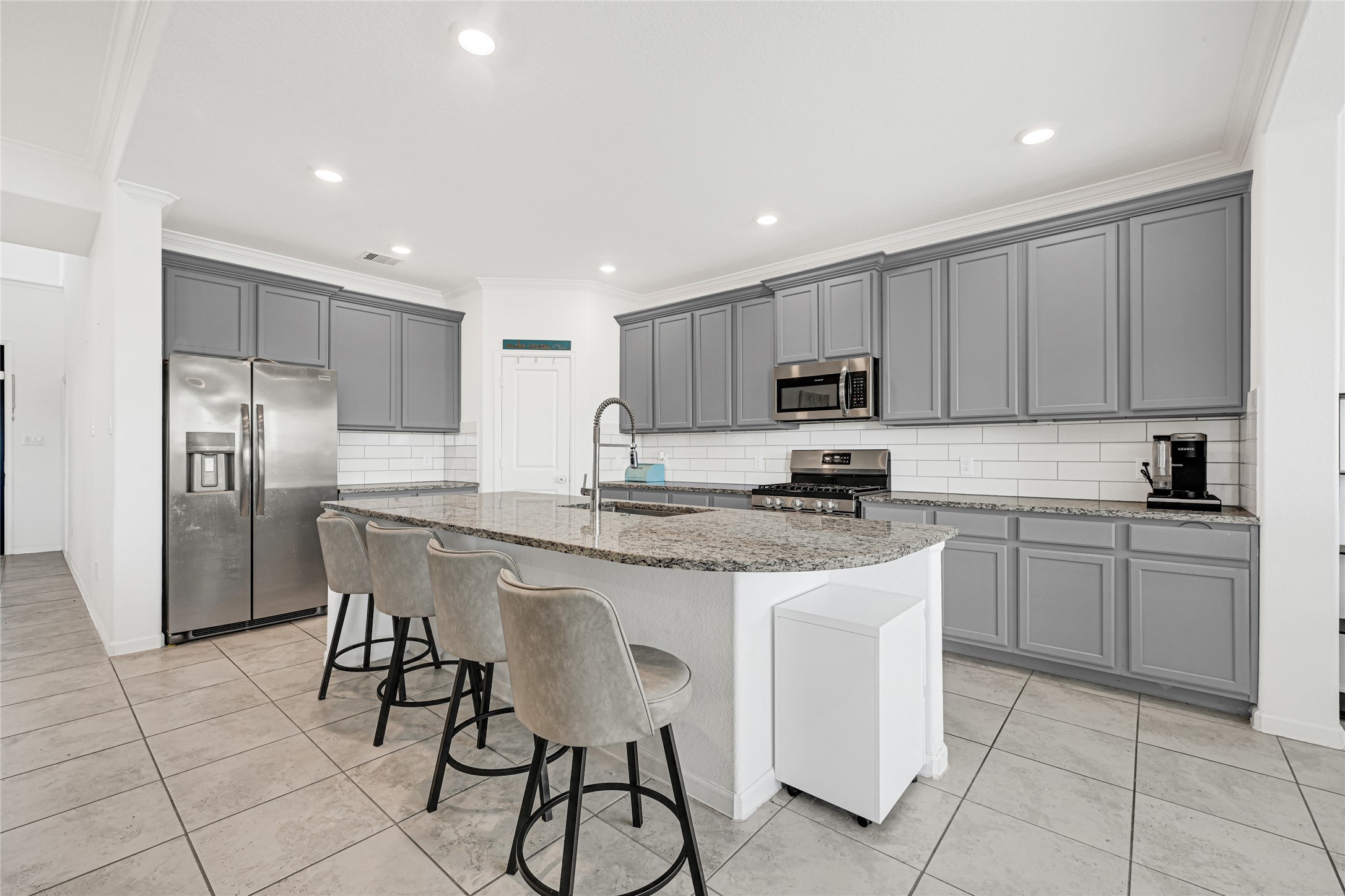 4307 Annandale Lane Rosharon, TX 77583 - Photo 3 of 31 This photo showcases a modern kitchen with gray cabinets, stainless steel appliances, and a large granite island with seating for four. The space is bright, featuring tiled floors and recessed lighting, creating an ample inviting and functional cooking area.