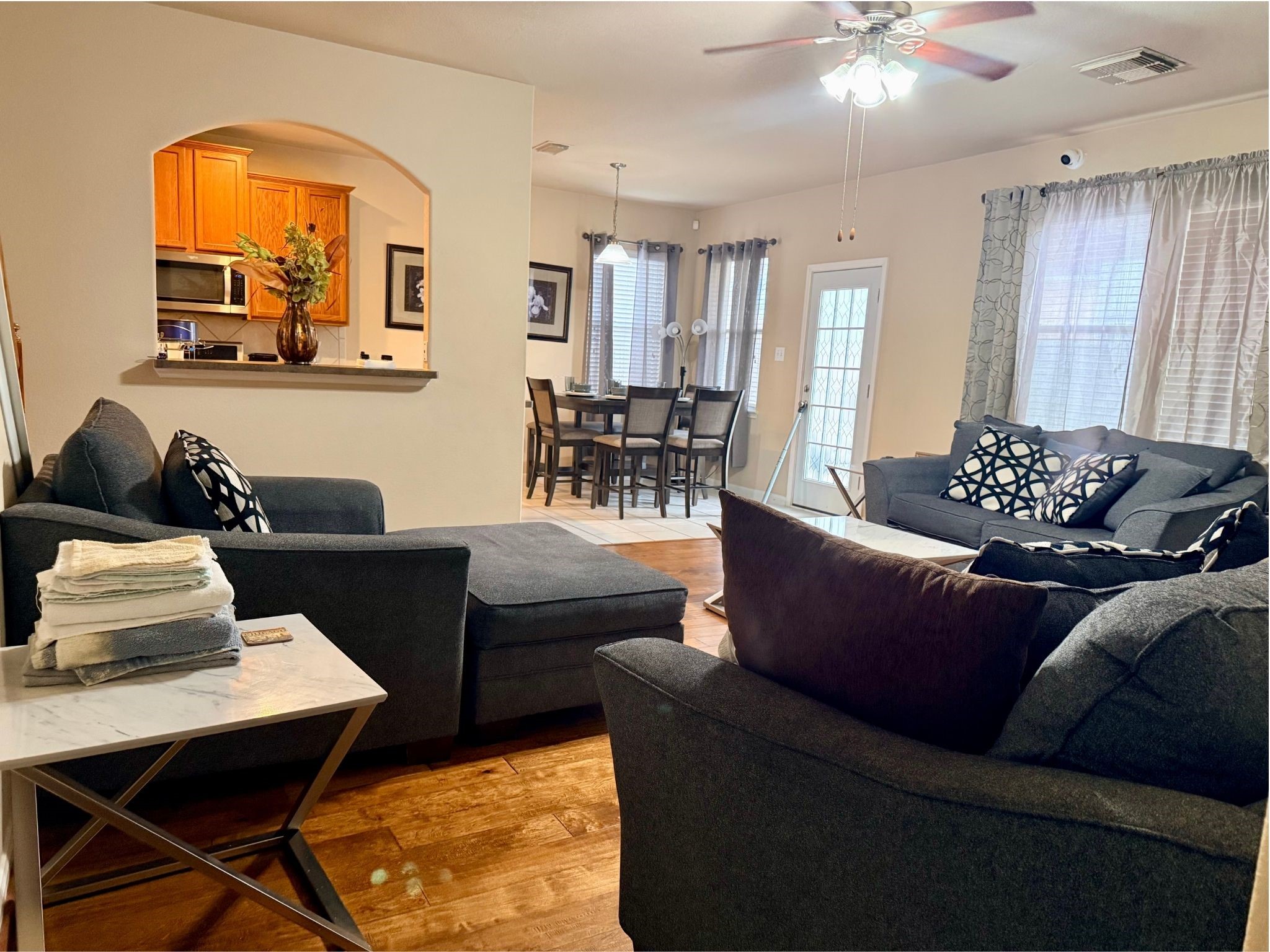 21831 Catoosa Drive Spring, TX 77388 - Photo 2 of 12 This inviting open-concept living space features a cozy seating area with plush gray sofas, a modern kitchen with warm wood cabinets visible through an arched opening, and a dining area with plenty of natural light. Hardwood floors and stylish decor complete the look.