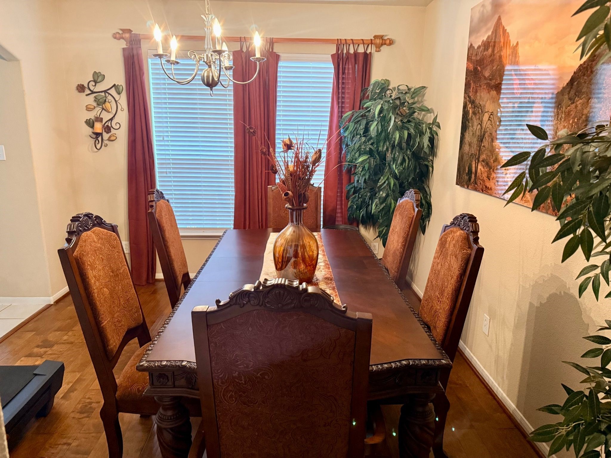 21831 Catoosa Drive Spring, TX 77388 - Photo 6 of 12 This photo shows a cozy dining room with a wooden table and six upholstered chairs. The room features warm decor, including a chandelier, wall art, and plants, creating an inviting atmosphere. Natural light filters through the large windows with red curtains, enhancing the space's elegance.