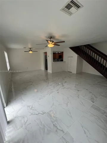 an empty room with chandelier fan and kitchen view