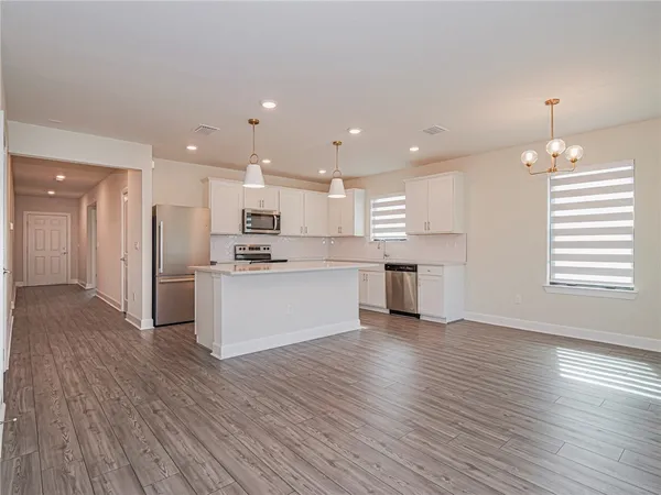 an empty room with chandelier fan and kitchen view