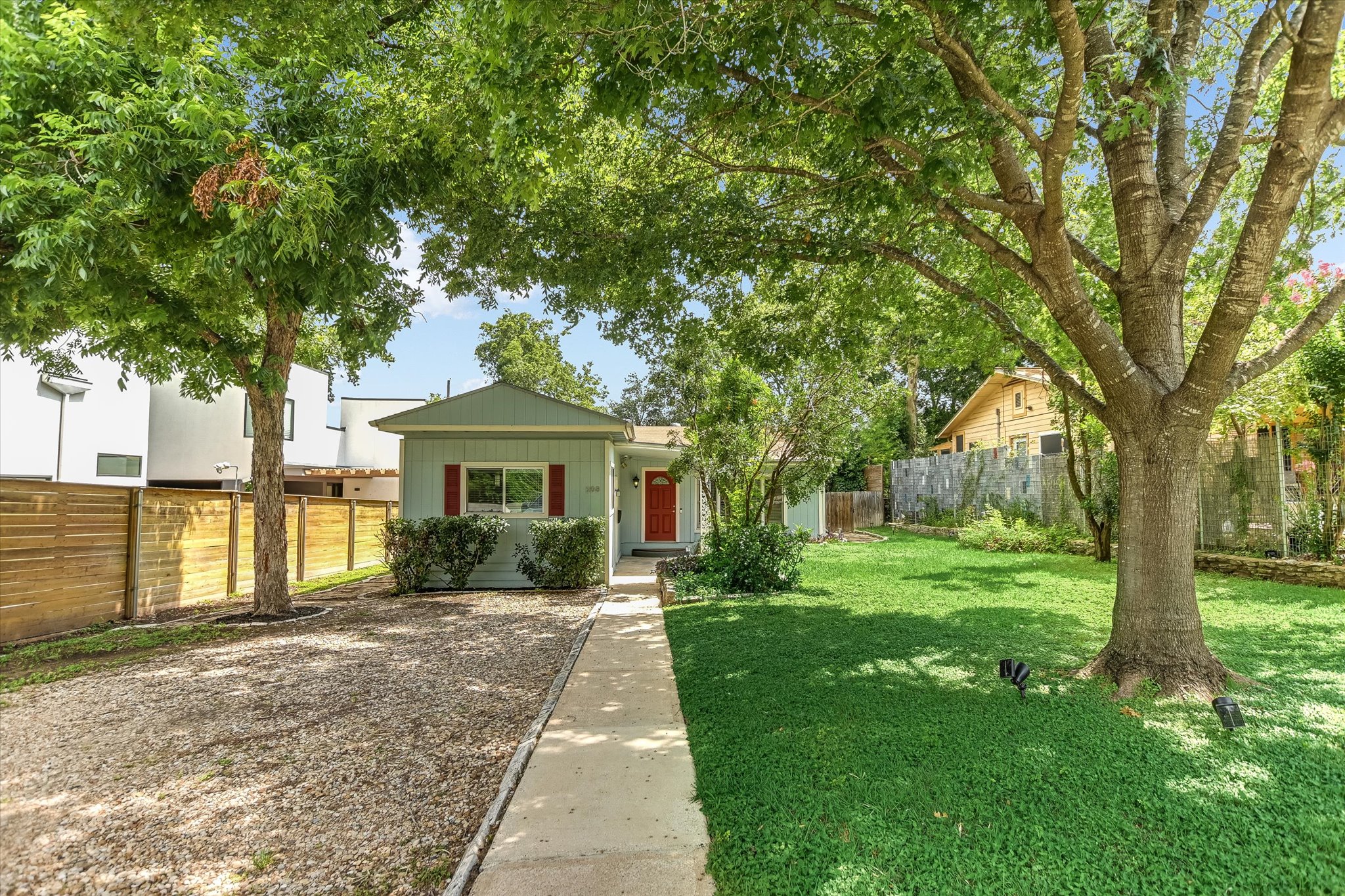 A welcoming light blue home with a red door is nestled amongst lush trees, boasting a well-maintained lawn and a stone walkway.