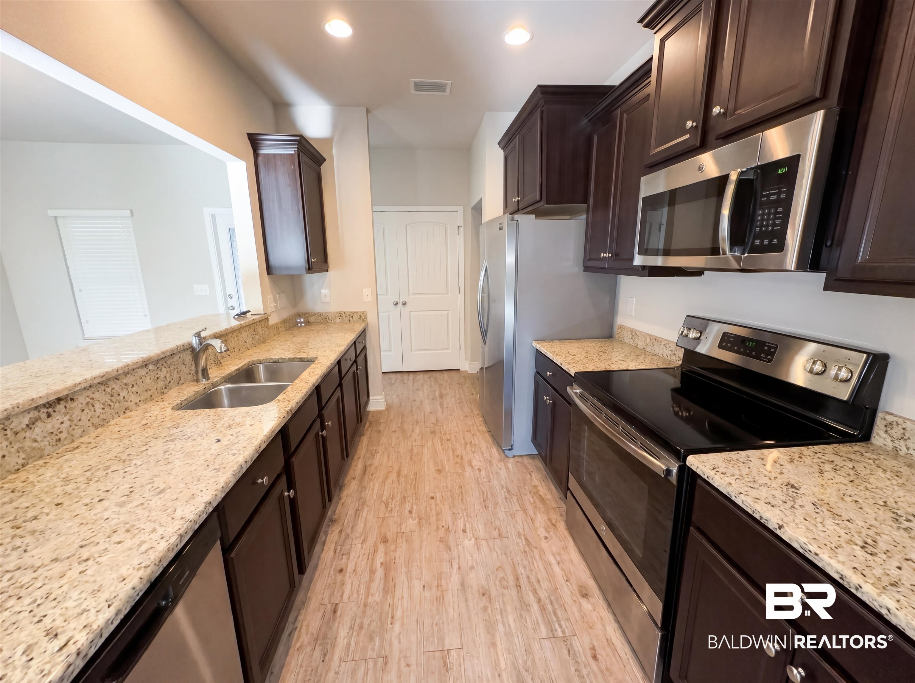 1740 Covington Lane Foley, AL 36535 - Photo 6 of 18 Galley kitchen featuring wood-finish flooring, dark wood cabinetry, light granite countertops, stainless steel appliances, and a pass-through window