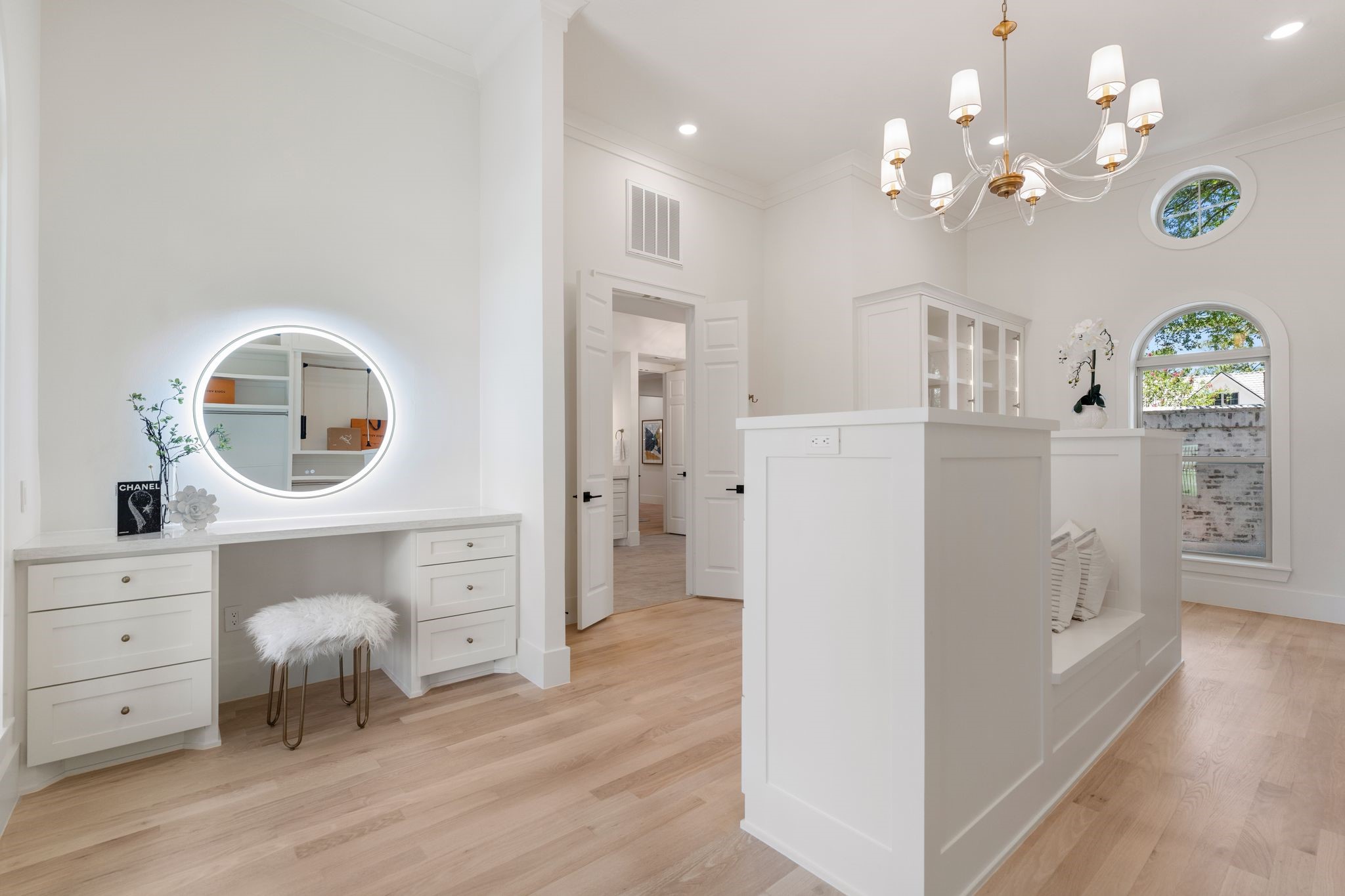 31 Hollymead Drive Spring, TX 77381 - Photo 27 of 50 This photo showcases a bright, elegant room with light wood flooring and white walls. It features a built-in vanity with a round illuminated mirror, a cozy seating nook, and stylish cabinetry. A modern chandelier and unique circular windows add a touch of sophistication.
