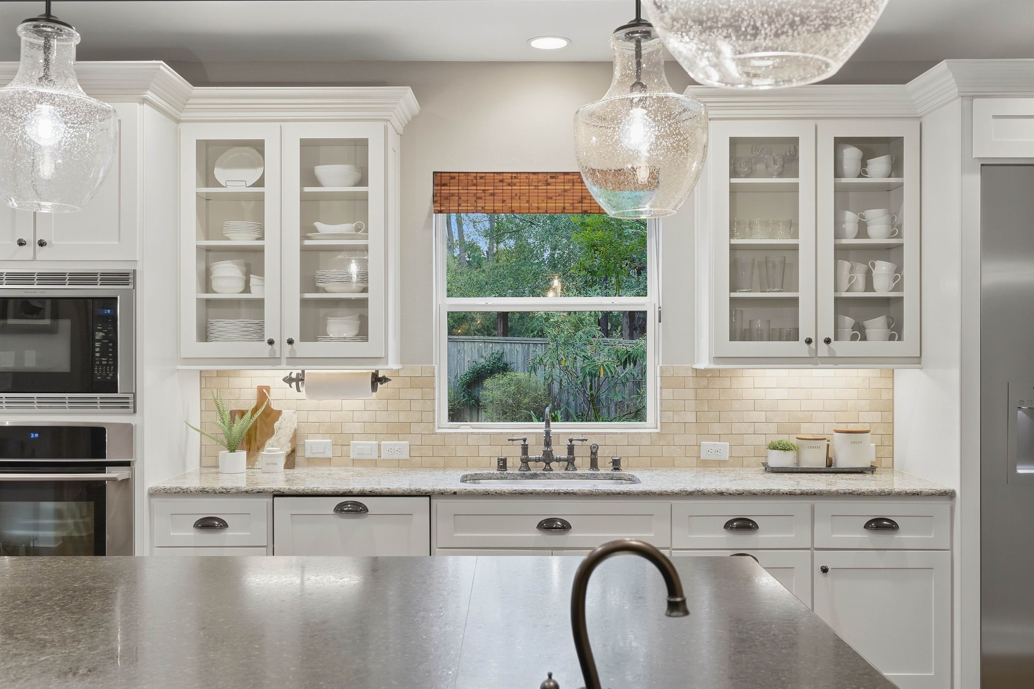 147 West Valera Ridge Place The Woodlands, TX 77389 - Photo 15 of 50 Bright kitchen with white cabinetry, glass-front uppers, granite countertops, classic subway tile backsplash, and a window overlooking the large side yard of the home.