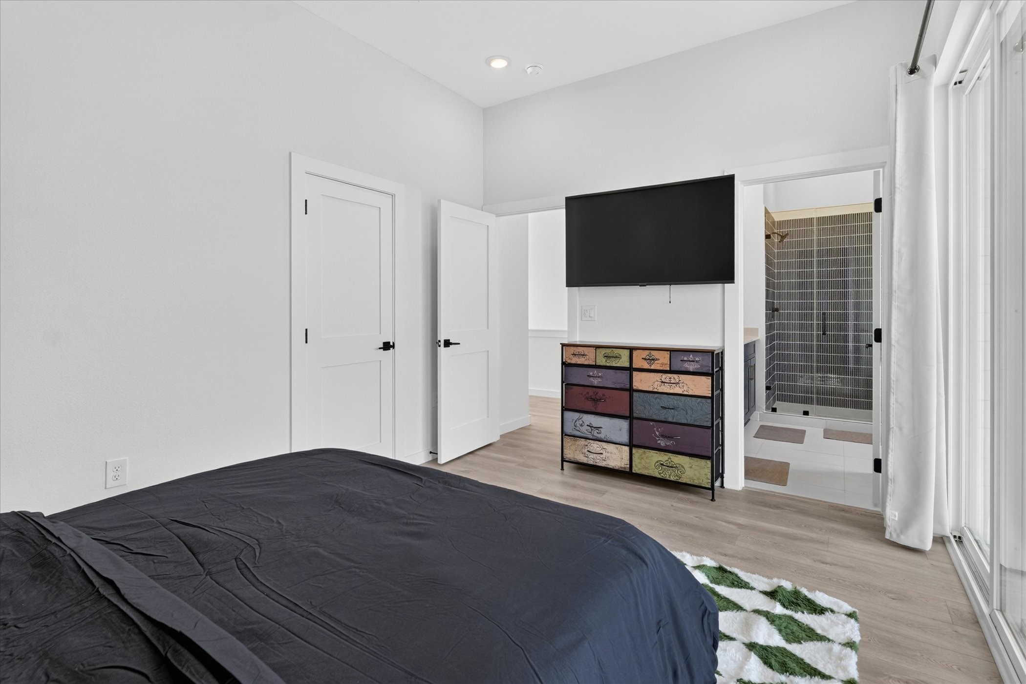 2000 Riverside Farms Road, Unit 8101 Austin, TX 78741 - Photo 22 of 41 This bedroom features a modern, clean design with light wood flooring and a neutral color palette. It includes a mounted TV above a colorful dresser, access to a private bathroom with a glass shower, and ample natural light from large windows.