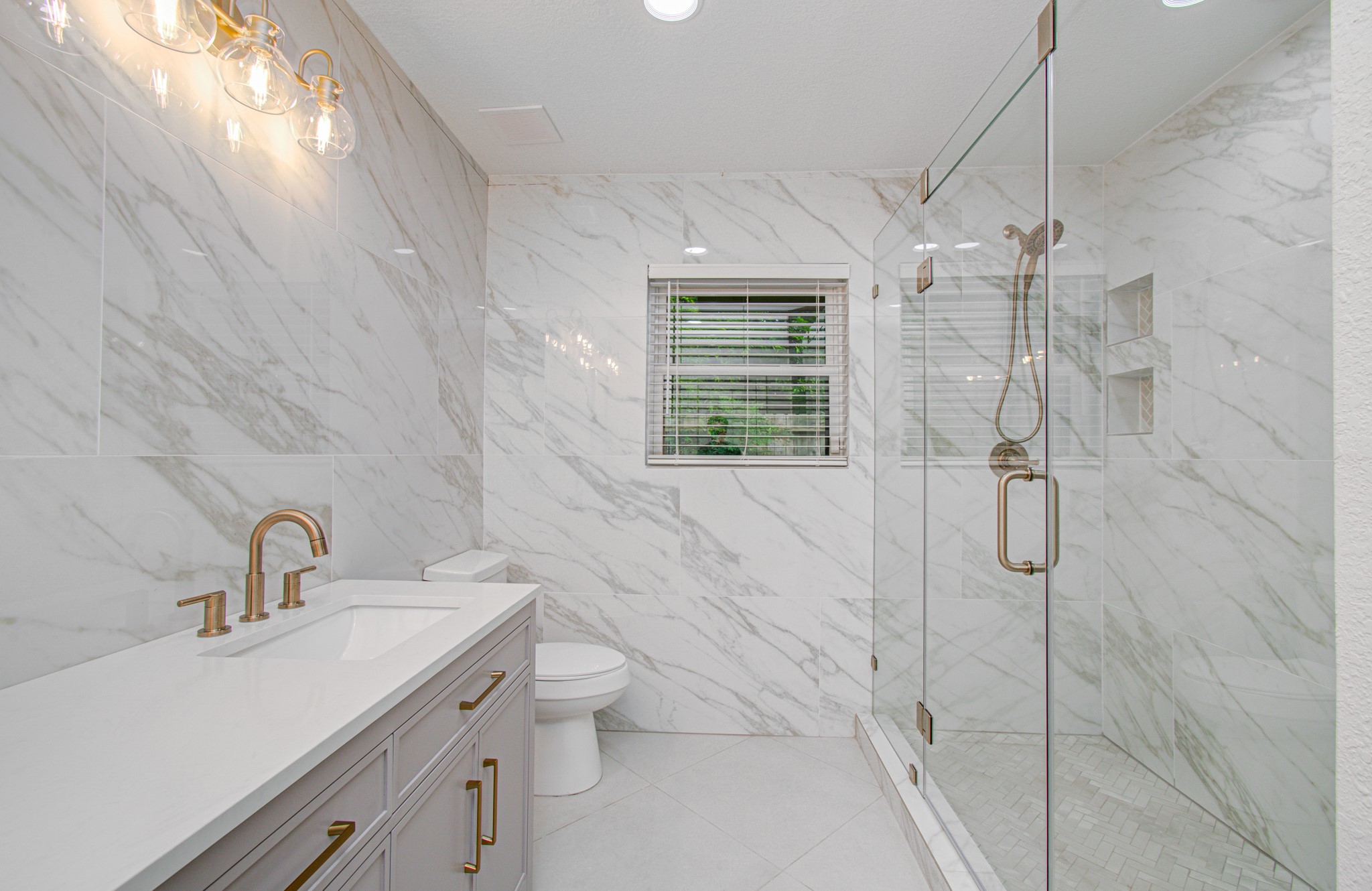 1801 Benbow Way Houston, TX 77080 - Photo 23 of 32 This bathroom features elegant white marble walls and flooring, a sleek vanity with gold fixtures, and a spacious glass-enclosed shower, creating a luxurious and modern feel.