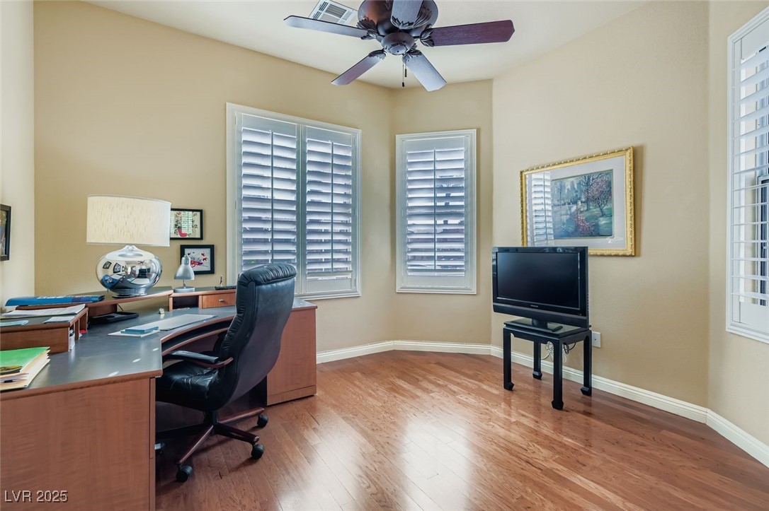 5015 Rivedro Street Las Vegas, NV 89135 - Photo 12 of 63 This home office features light wood-style flooring, elegant plantation shutters, and a sleek ceiling fan—all bathed in abundant natural light. A perfect blend of comfort and productivity in a beautifully appointed space.
