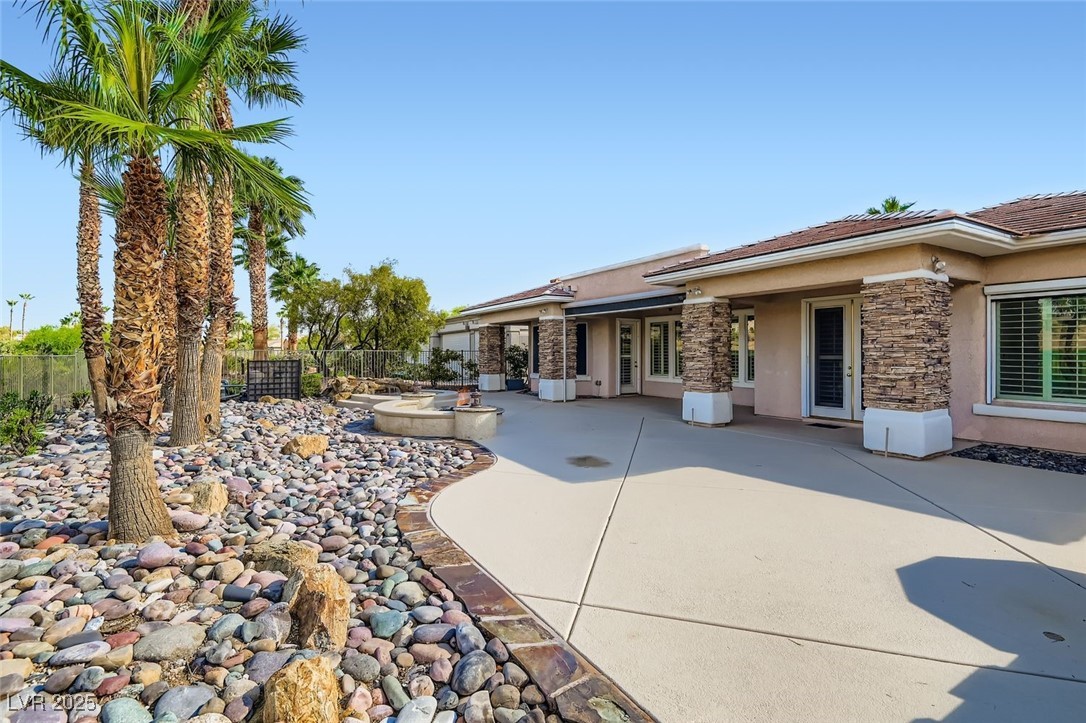 5015 Rivedro Street Las Vegas, NV 89135 - Photo 13 of 63 From the southwest corner of this spacious lot, the image captures lush, meticulously maintained landscaping, stone-trimmed pillars that frame the patio with timeless character, and the French door entry into the main living space—blending indoor comfort with outdoor sophistication.