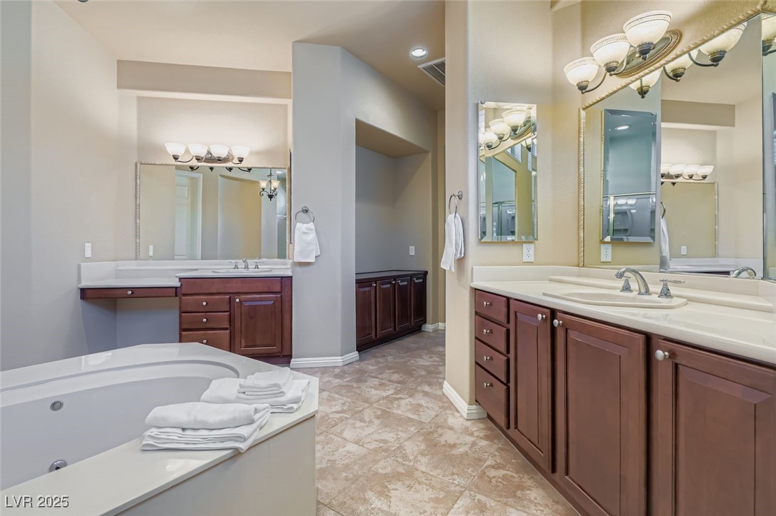 5015 Rivedro Street Las Vegas, NV 89135 - Photo 9 of 63 This view of the primary bath showcases dual vanities, a relaxing whirlpool tub, and light tile-patterned flooring illuminated by recessed lighting. Just beyond, additional countertop cabinetry in the hallway offers extra storage and a seamless transition to the primary bedroom—blending beauty with practicality.