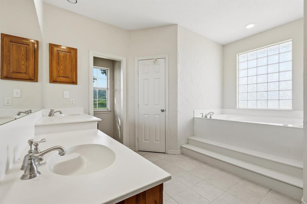 12190 Bell Road Pilot Point, TX 76258 - Photo 23 of 39 Spacious bathroom featuring a built-in soaking tub with tiered steps, a large glass block window, double vanity sinks with white countertops, and wood cabinetry