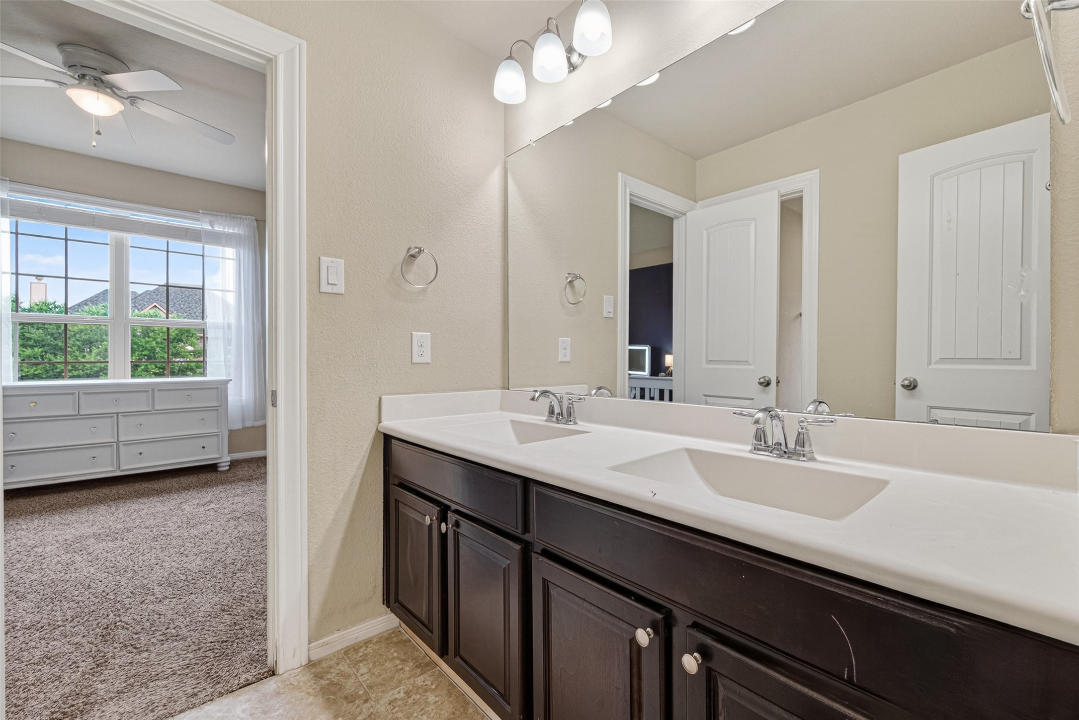 25611 Ellis Ridge Court Katy, TX 77494 - Photo 31 of 49 This photo shows a bright bathroom with dual sinks, dark wood cabinetry, and a large mirror. Adjacent is a carpeted bedroom with large windows, a ceiling fan, and a white dresser, offering a cozy and functional living space.