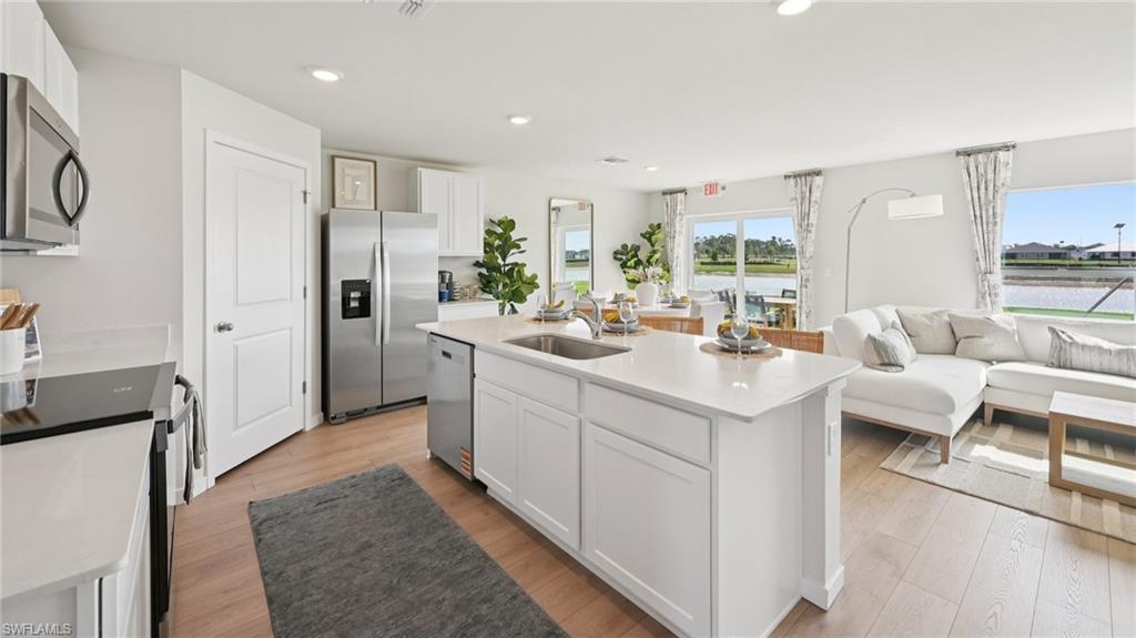 5007 3rd Street West Lehigh Acres, FL 33971 - Photo 7 of 24 Model Home. The open concept living area features light wood flooring, a kitchen island with a sink, white cabinetry, and stainless steel appliances