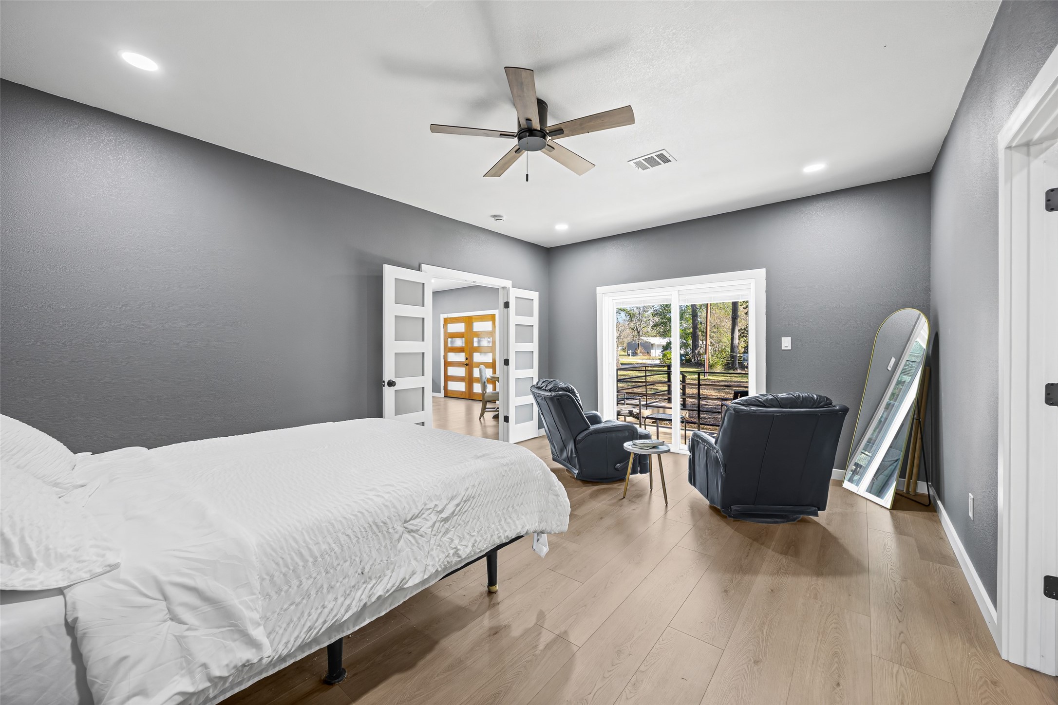 15080 Usher Lane Splendora, TX 77372 - Photo 24 of 44 This spacious bedroom features modern gray walls, light wood flooring, and a ceiling fan. It includes a cozy seating area with two chairs by large sliding glass doors that open to a view. Ample natural light and a minimalist design create a serene ambiance.