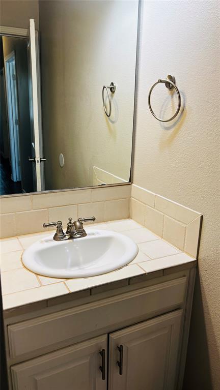 4511 Junius Street Dallas, TX 75246 - Photo 10 of 16 Bathroom 1 vanity featuring a tiled countertop and backsplash, undermount sink with brushed nickel faucet, full-width wall mirror, and lower cabinetry with storage