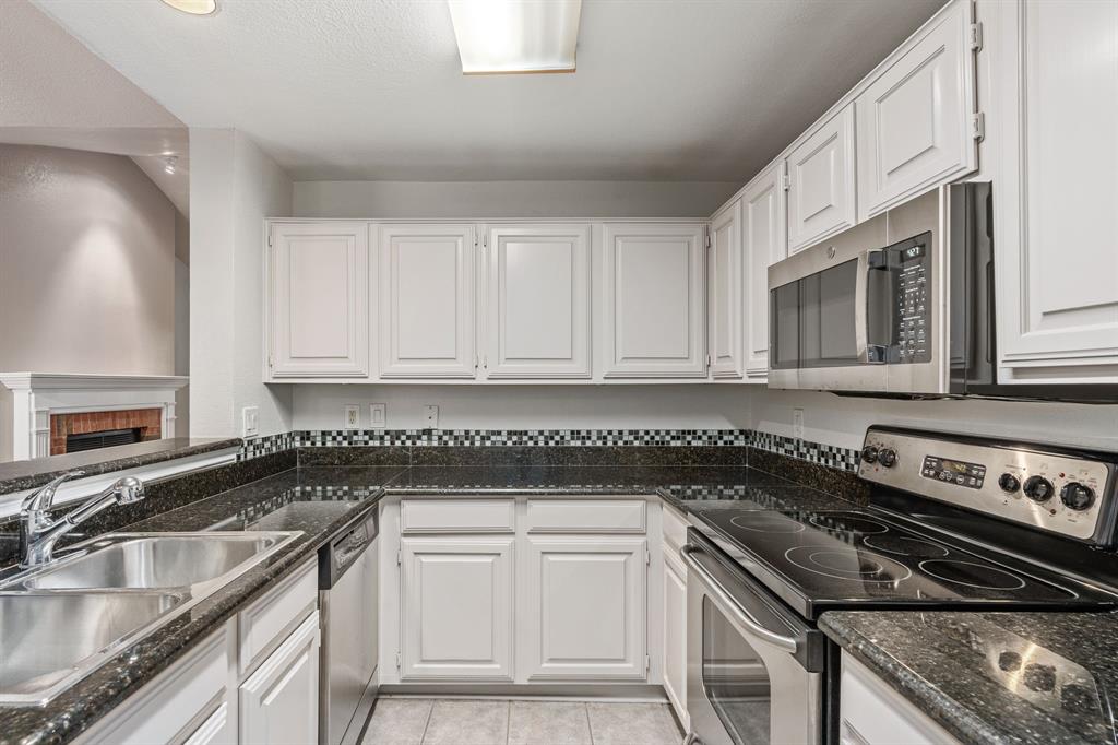 12660 Hillcrest Road, Unit 4203 Dallas, TX 75230 - Photo 7 of 16 Kitchen featuring a brick fireplace, dark stone counters, stainless steel appliances, light tile patterned floors, and white cabinets
