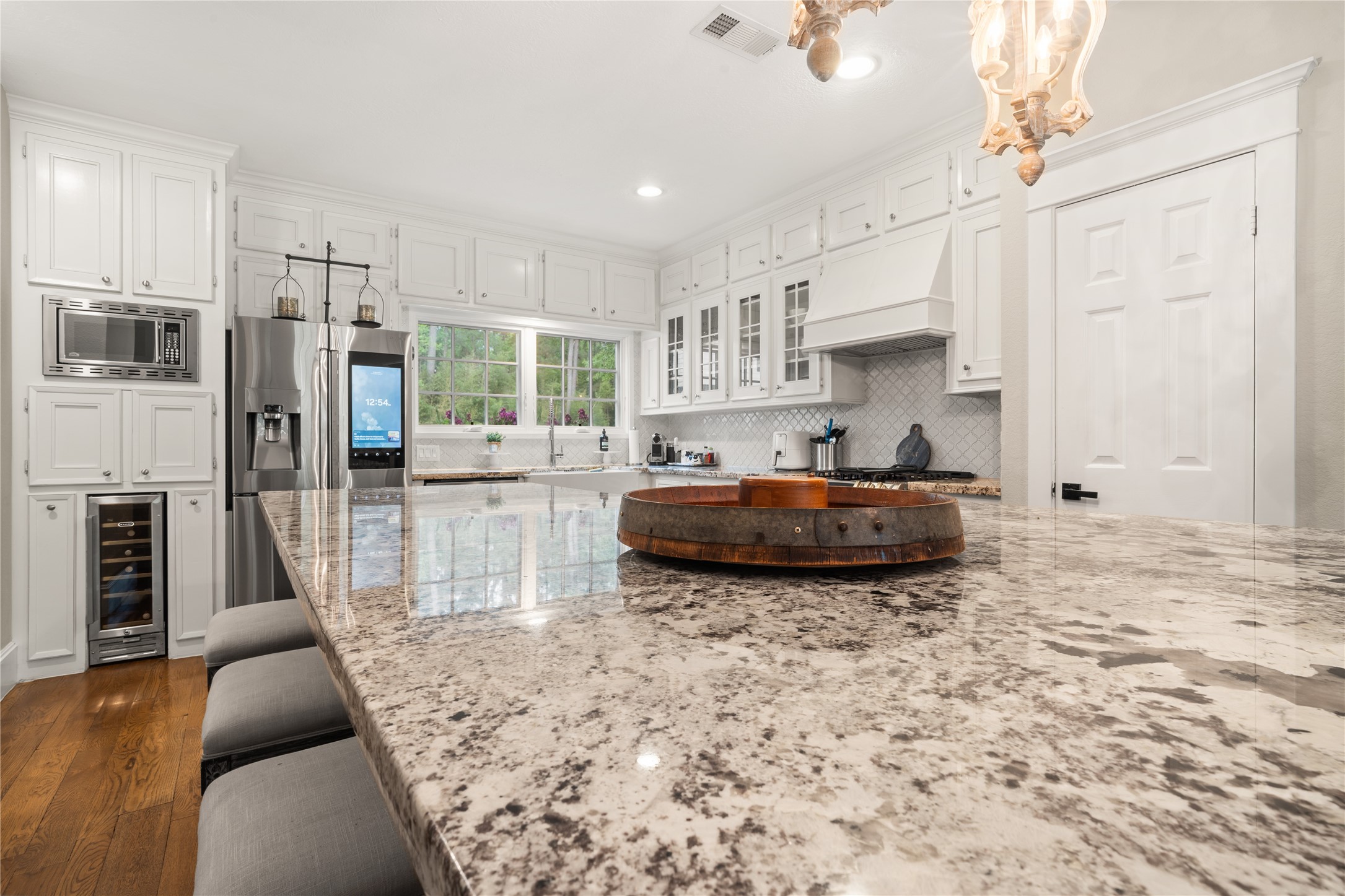 36 Wind River Drive Conroe, TX 77384 - Photo 22 of 45 This kitchen features a spacious granite island with seating, stainless steel appliances, and ample white cabinetry. It has a large window for natural light, hardwood floors, and elegant lighting fixtures, creating a bright and inviting space.