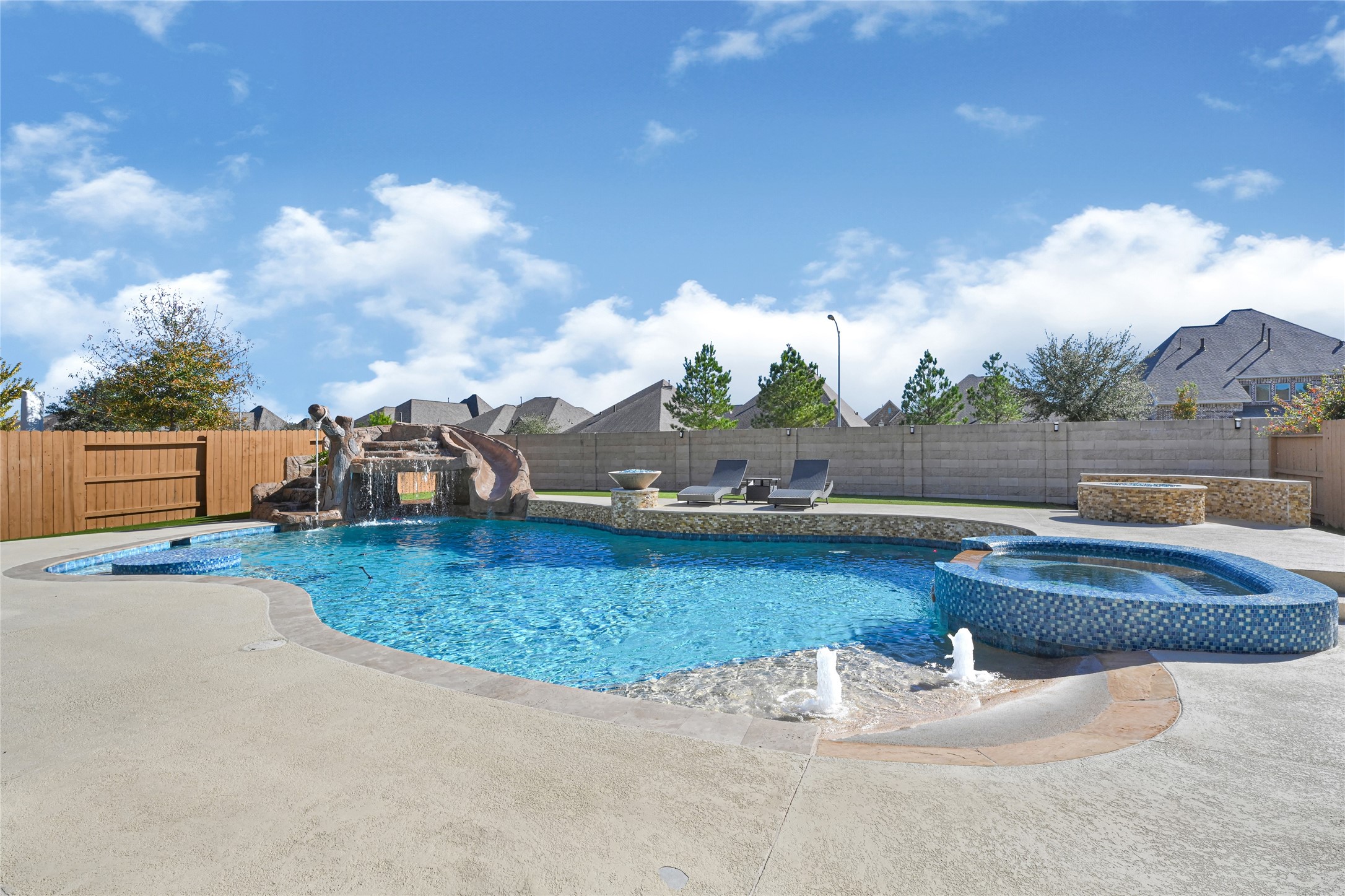 This backyard features a spacious pool with a water slide and a cascading waterfall. It includes a hot tub, lounge chairs, and a stone-accented seating area, perfect for relaxation and entertainment.